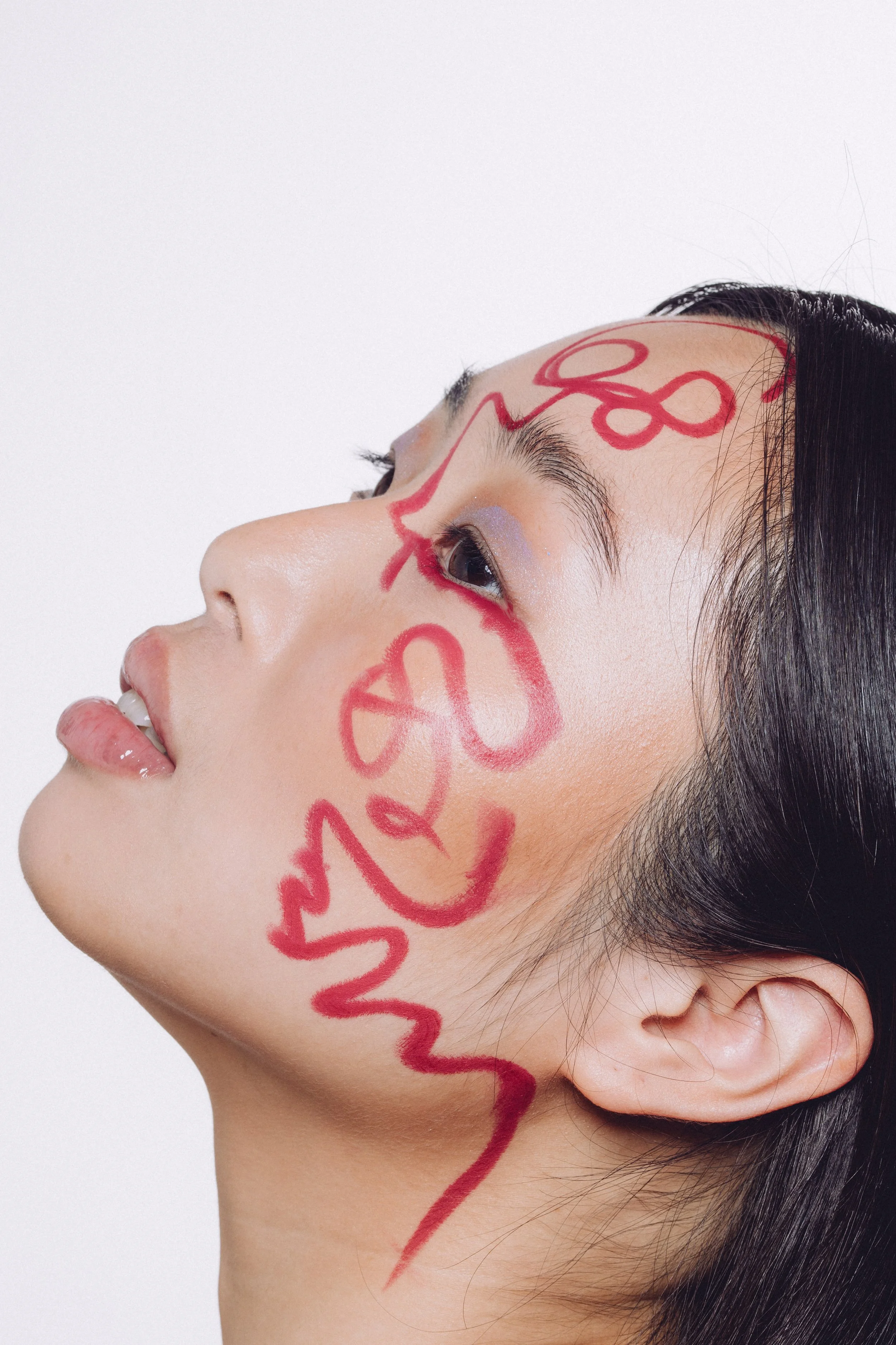 A woman with long black hair has the word "LOVE" written in red lipstick across her face, including her forehead, cheeks, and chin.