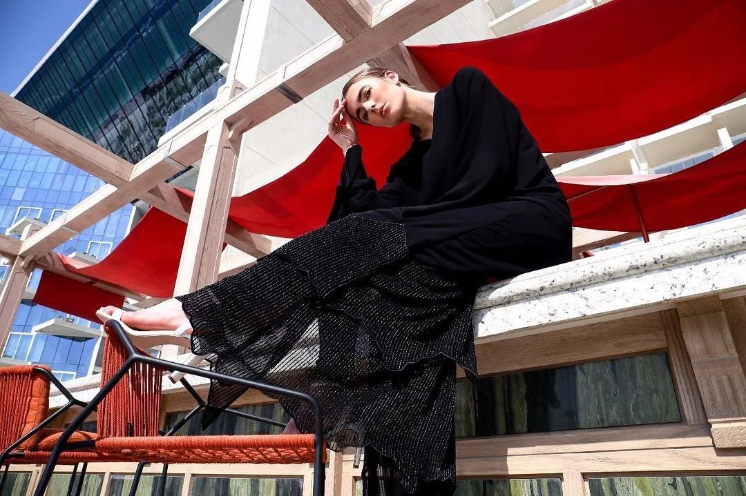A woman wearing black clothing is sitting on a concrete ledge under a wooden pergola with red fabric shades, outdoors in a city setting with tall glass buildings in the background.
