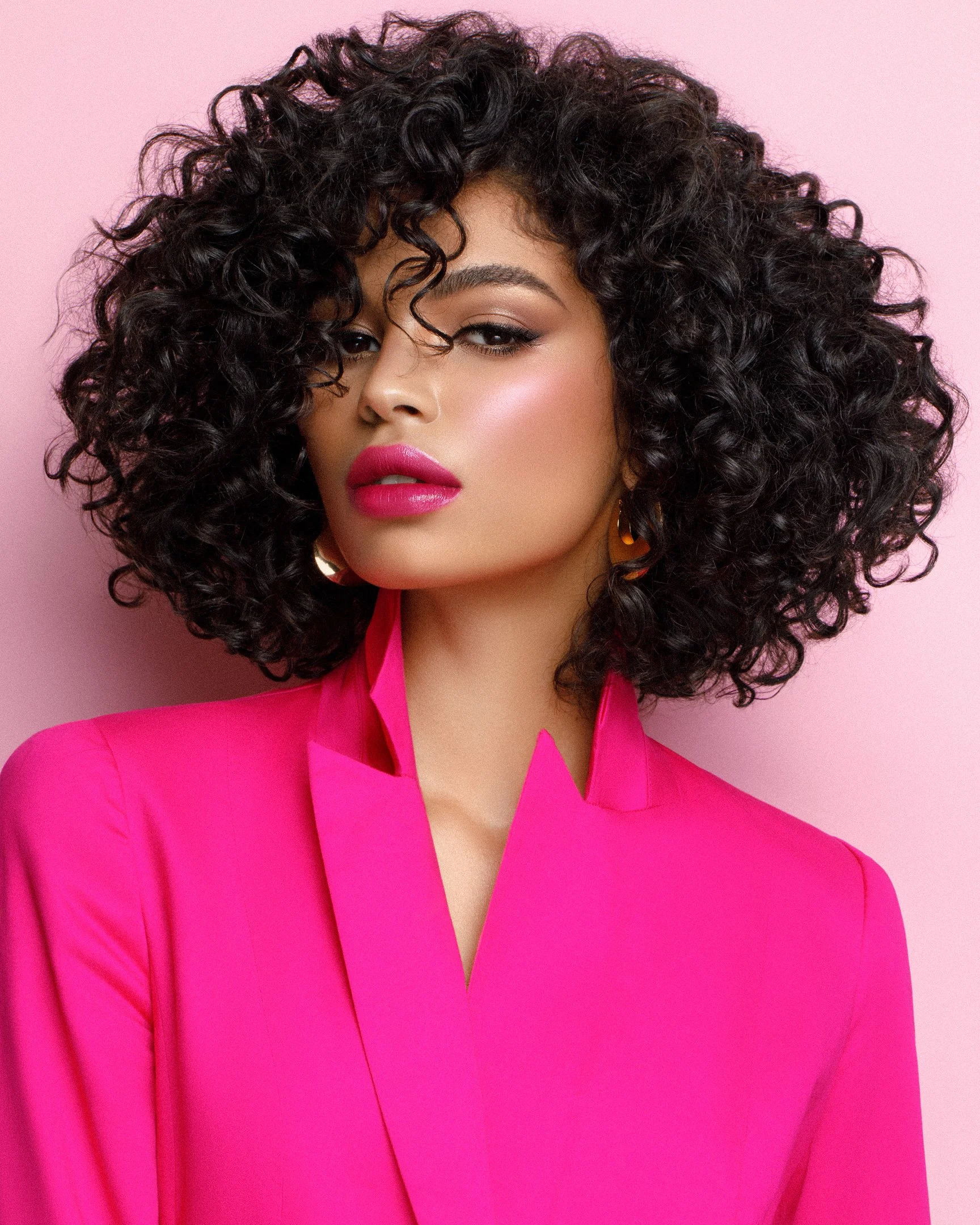 A woman with dark curly hair, wearing bright pink lipstick and matching pink blazer, against a pink background.