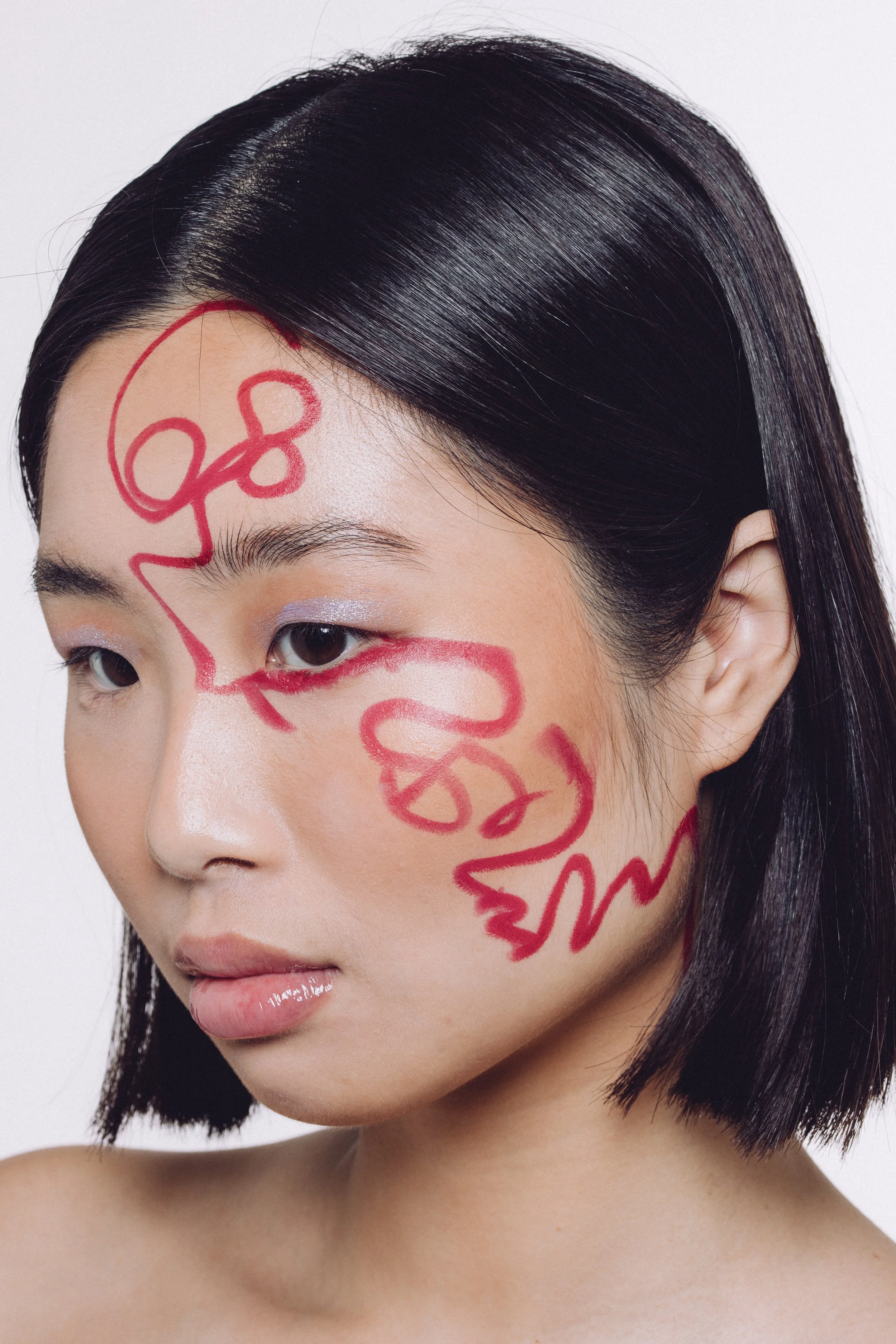 A woman with short black hair styled in a sleek bob, has red abstract line art on her face, with lavender eyeshadow on her eyelids.