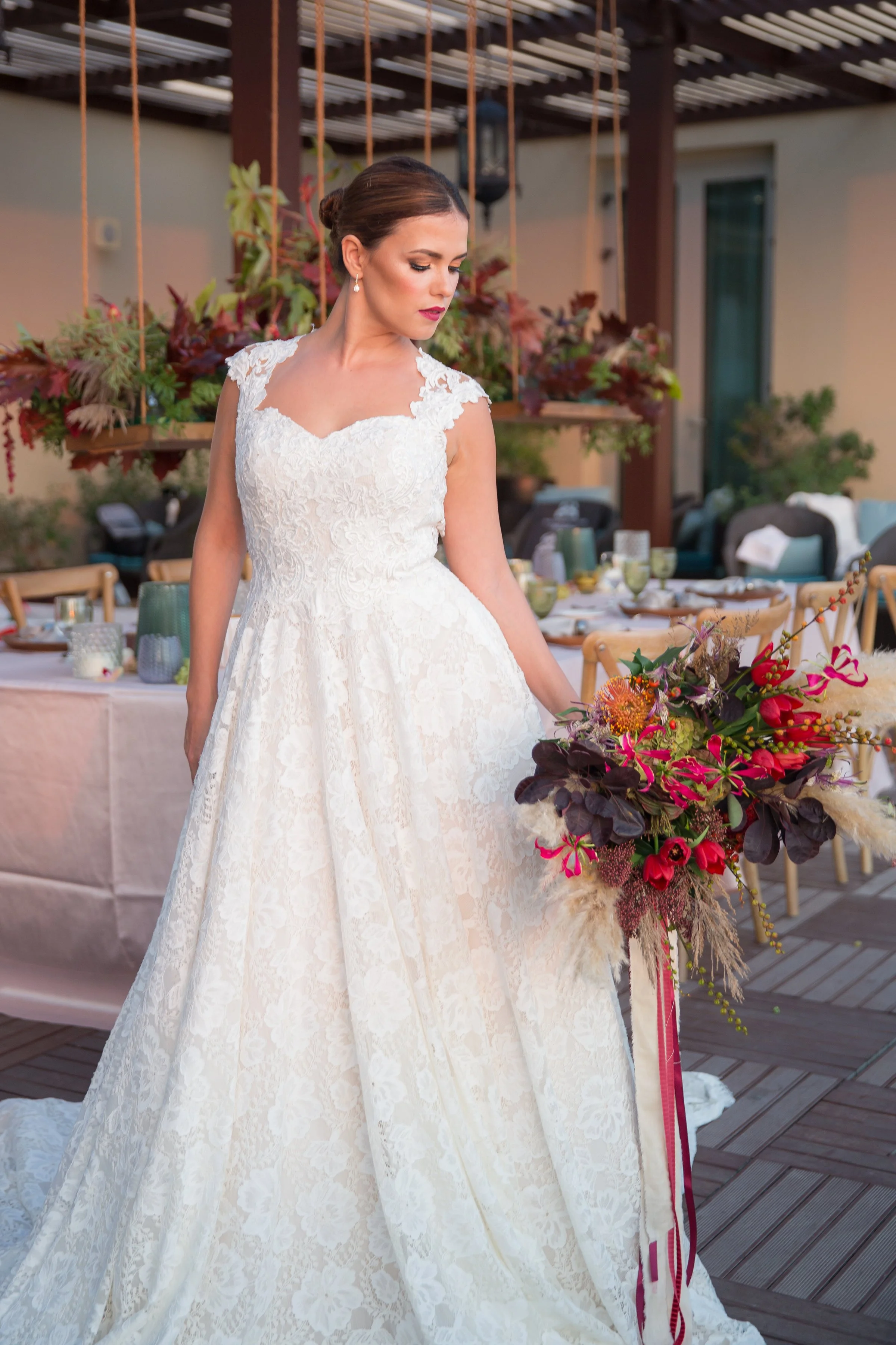 A bride in a white lace wedding gown holding a bouquet of red, pink, and dark purple flowers during a wedding reception outdoors.