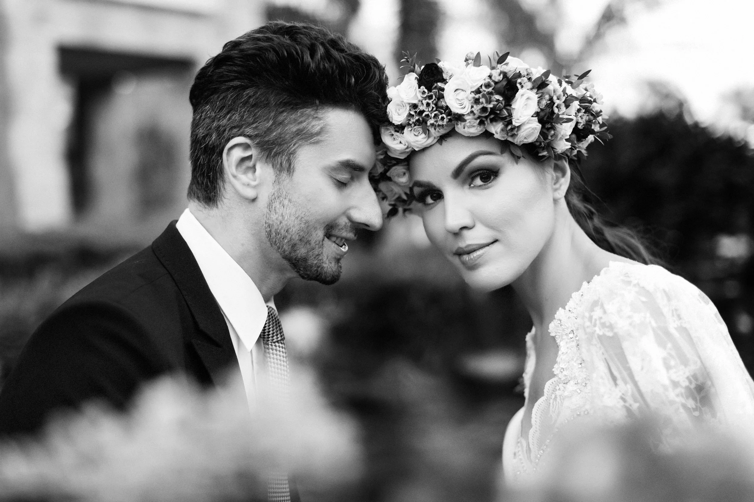 Black and white photograph of a bride and groom, with the groom leaning in and the bride looking at the camera, wearing a flower crown, outdoors.
