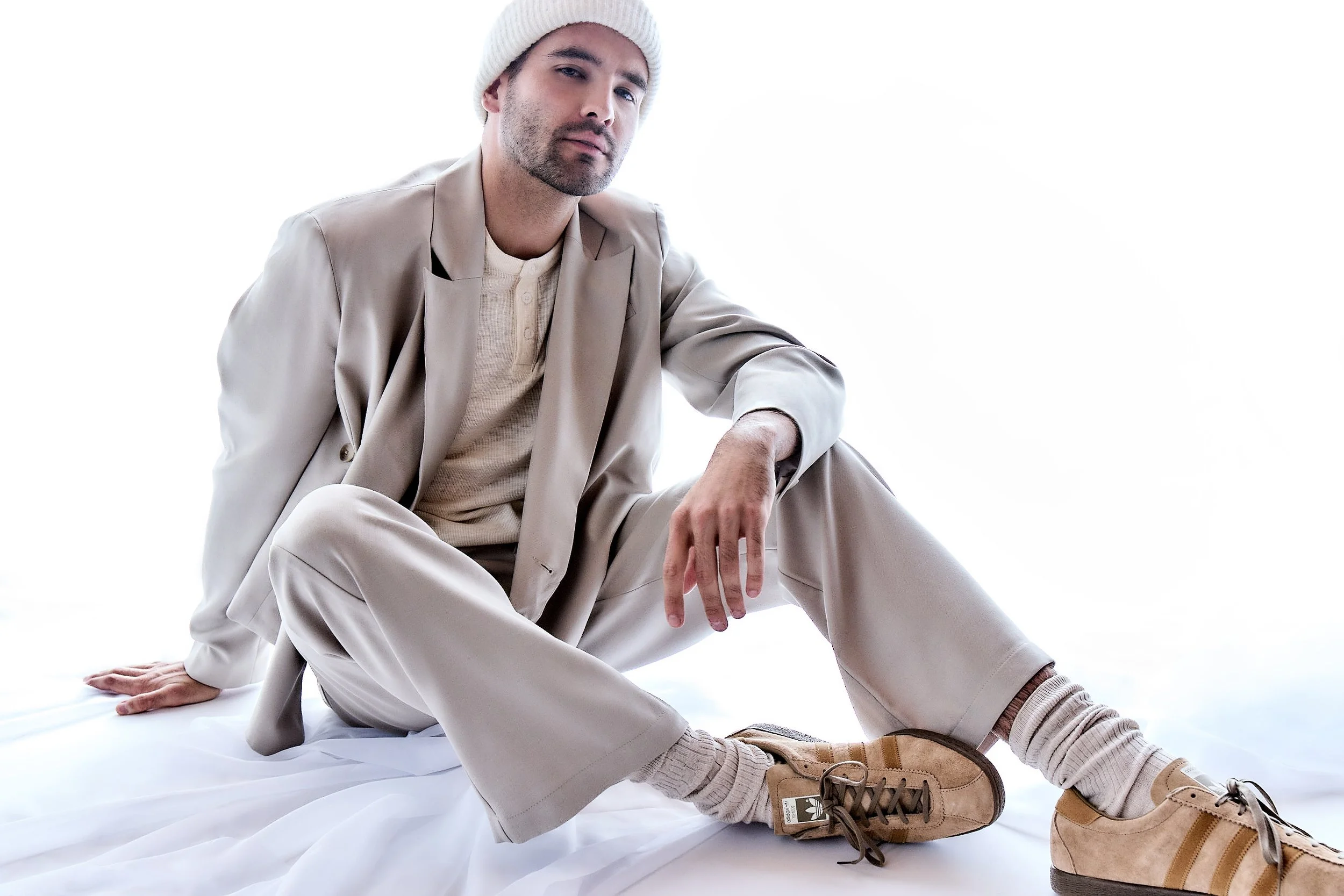 A man sitting on a white surface, dressed in beige clothing and sneakers, wearing a white beanie, looking at the camera with a relaxed pose.
