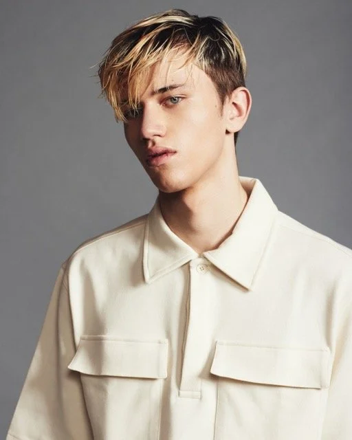 Young man with tousled blond hair wearing a cream-colored button-up shirt against a gray background.