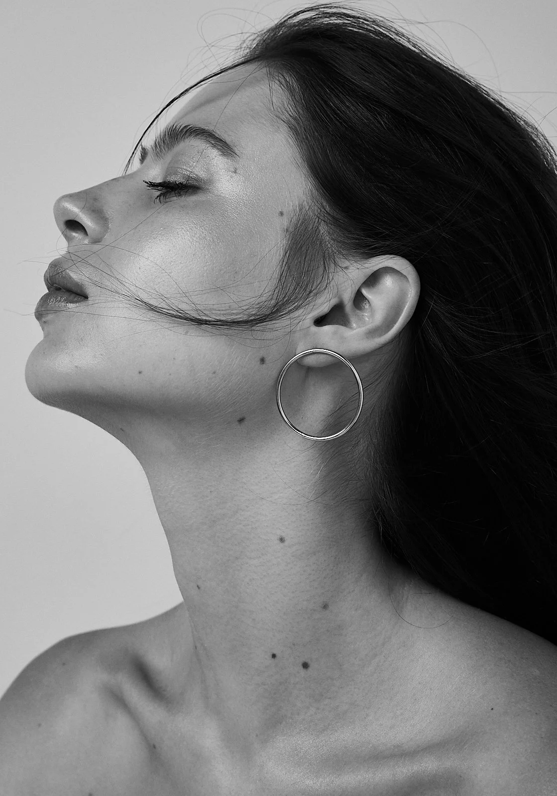 Black and white close-up photo of a woman with eyes closed, wearing a hoop earring, with her hair blowing across her face and neck.