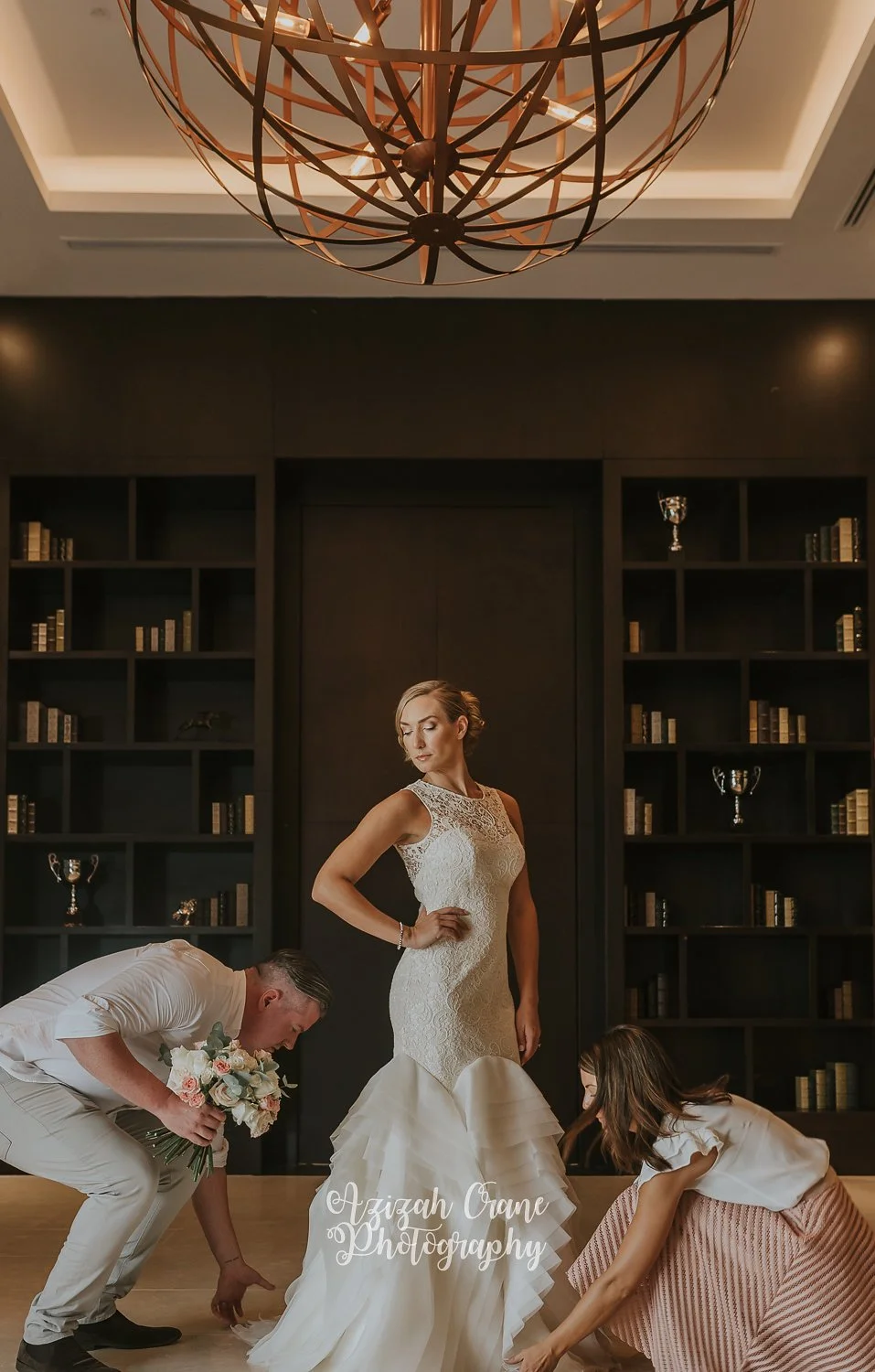A bride in a white lace wedding dress stands confidently while two women adjust her dress on either side. One woman is holding a bouquet, and the other is crouching down. The background features black shelves with books and trophies, and a decorative