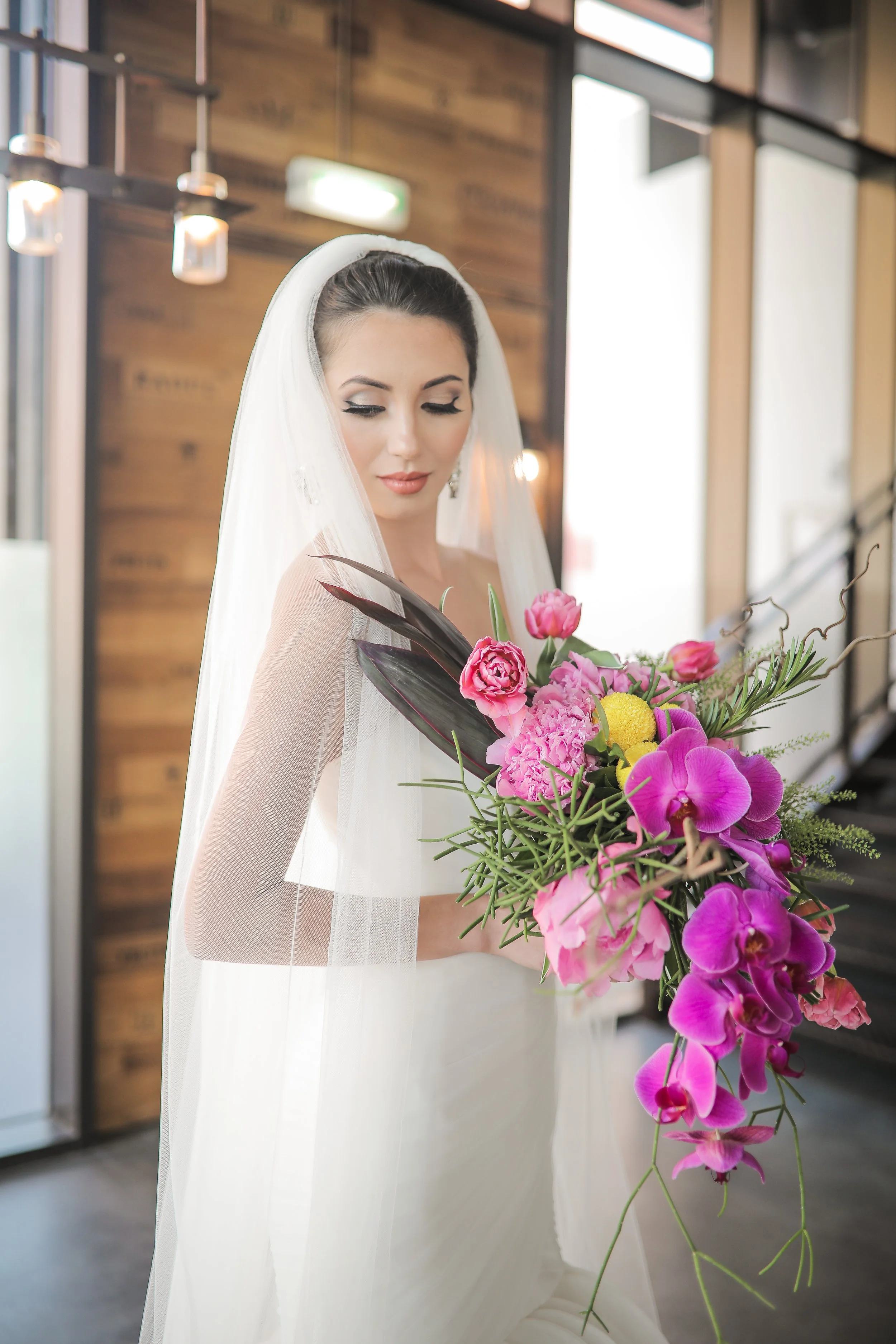 A bride wearing a white wedding gown and veil holding a vibrant bouquet of pink, purple, and yellow flowers inside a modern building with wood and glass decor.