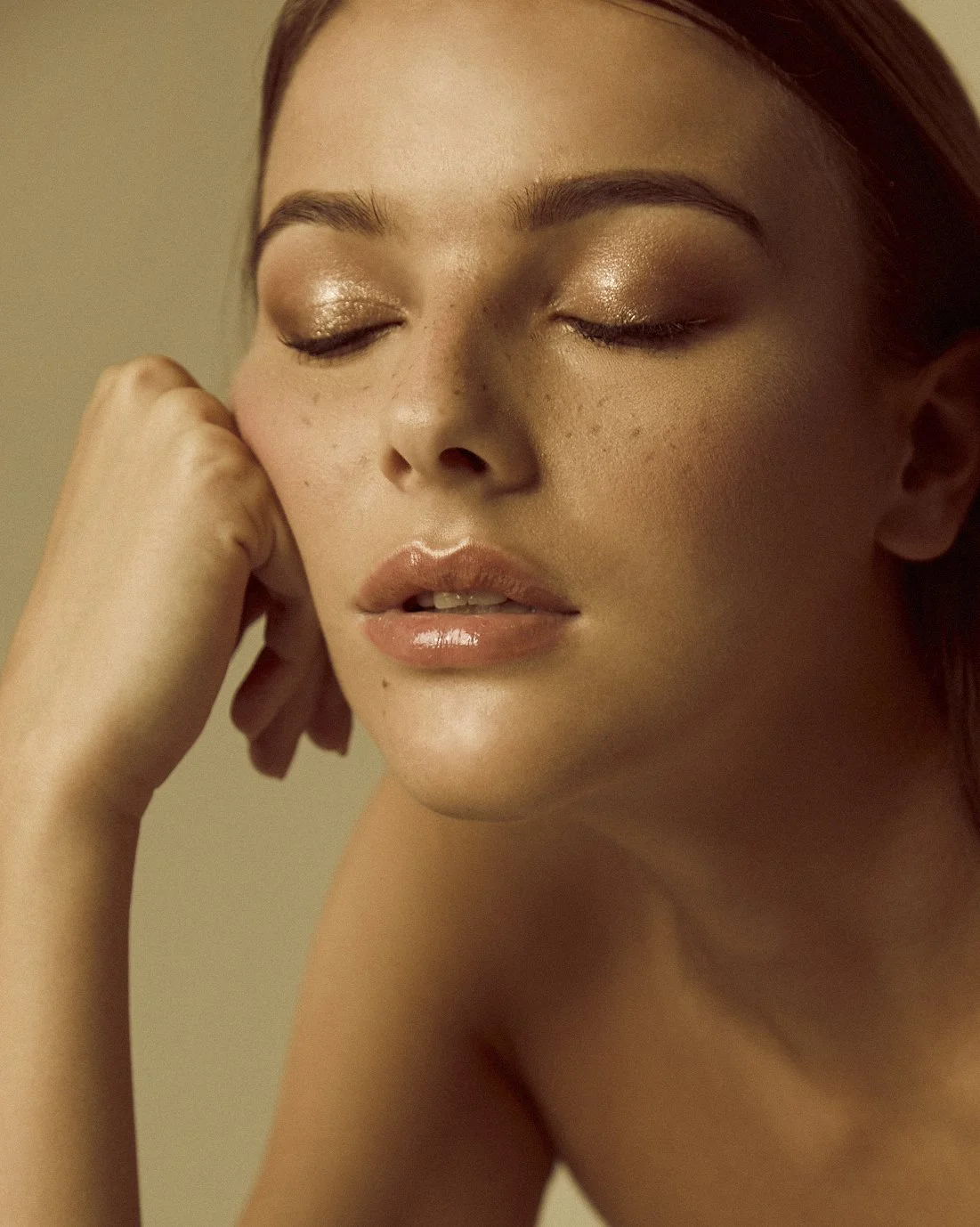 Close-up of a woman with closed eyes, warm makeup, and light skin, resting her face on her hand.