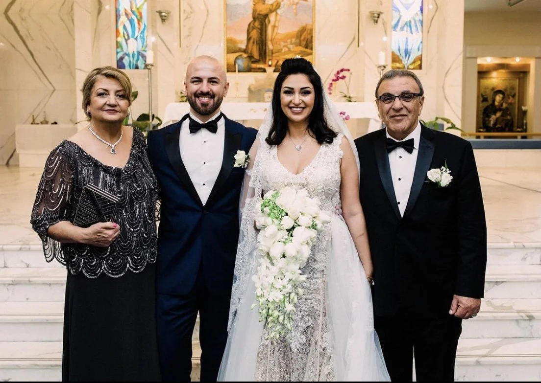 A wedding scene with four people standing inside a church. The bride is wearing a white wedding gown and holding a bouquet. The groom is dressed in a navy tuxedo with a bow tie. The woman on the far left is in a black dress with silver embellishments