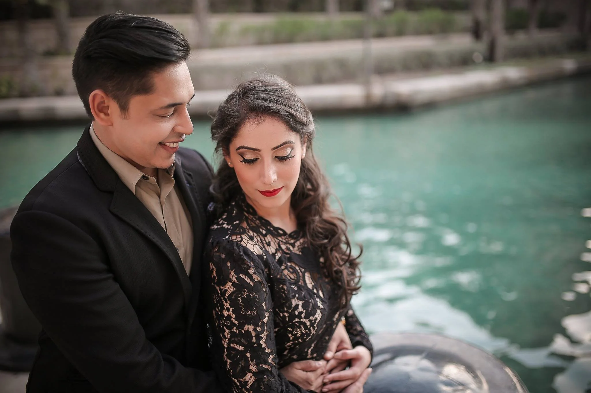 A young man and woman sit close by a body of water, with the man smiling while looking at the woman, who looks down. The woman is wearing a black lace dress, and the man is dressed in a black jacket and beige shirt.