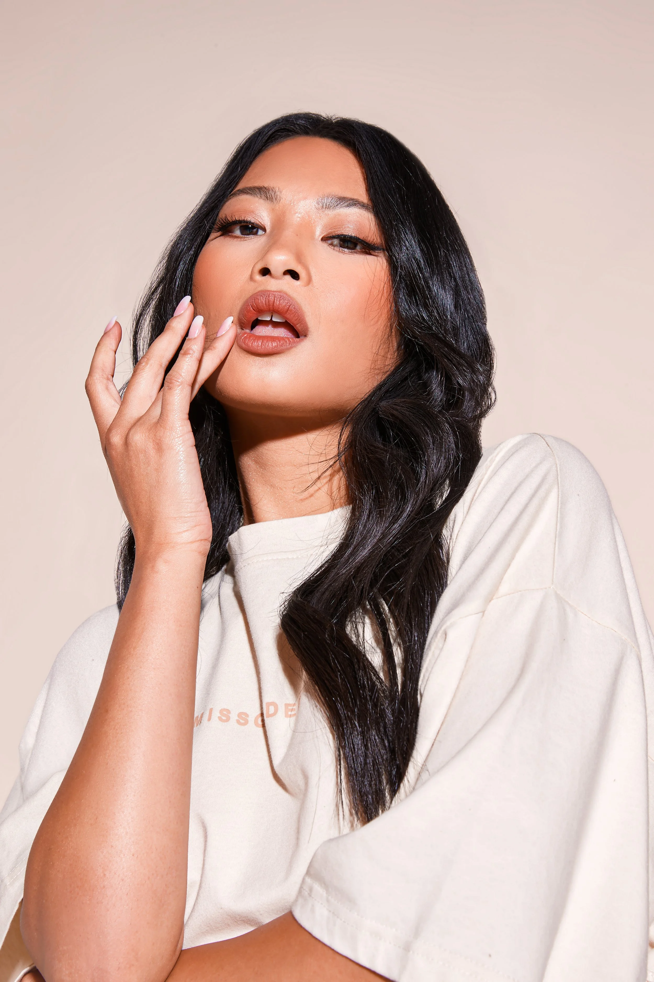 A woman with long dark wavy hair, wearing a light-colored T-shirt, posing with her hand near her face against a plain background.