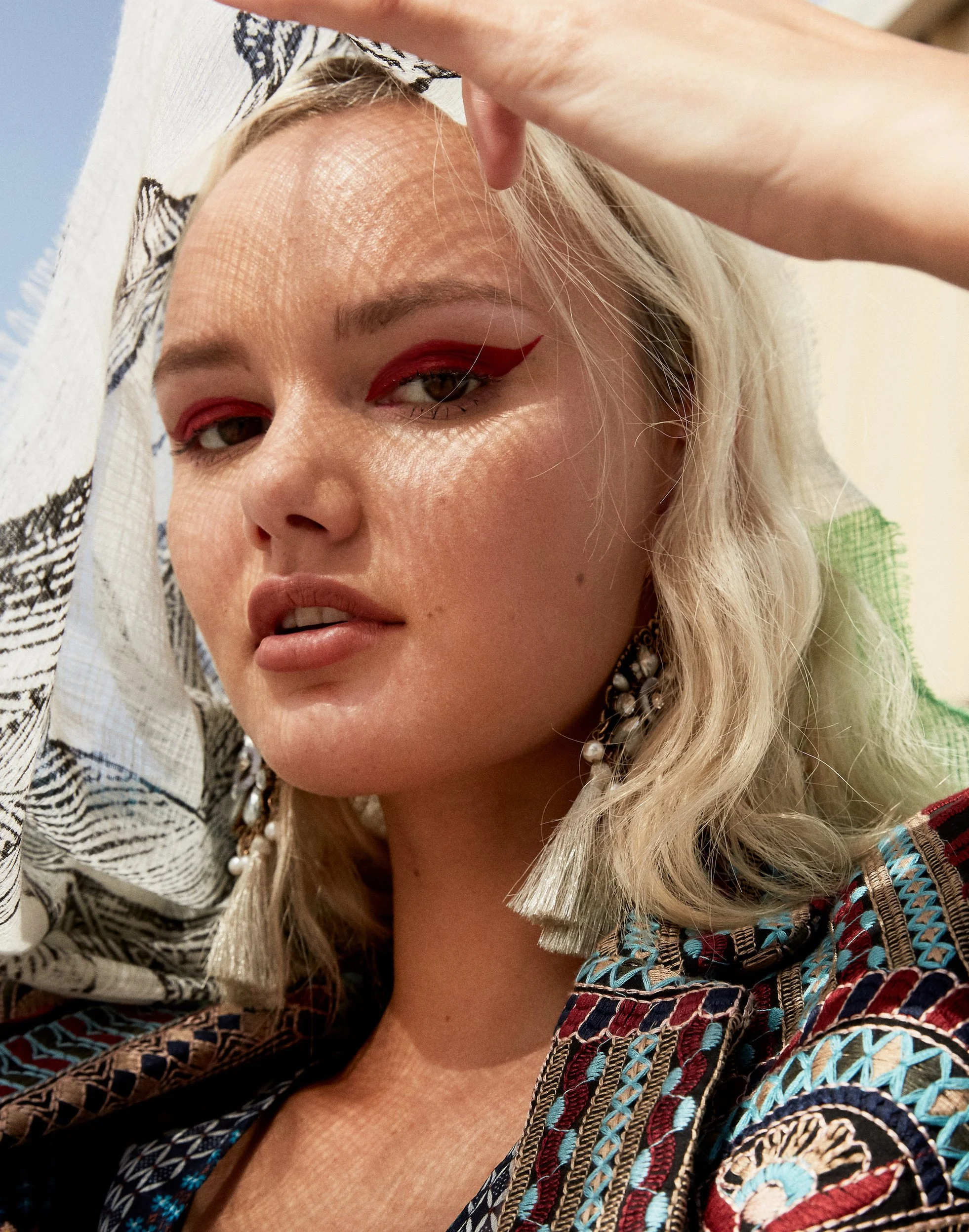 A woman with blonde hair is taking a close-up selfie. She has on bold red eyeliner, large earrings, and a patterned jacket.