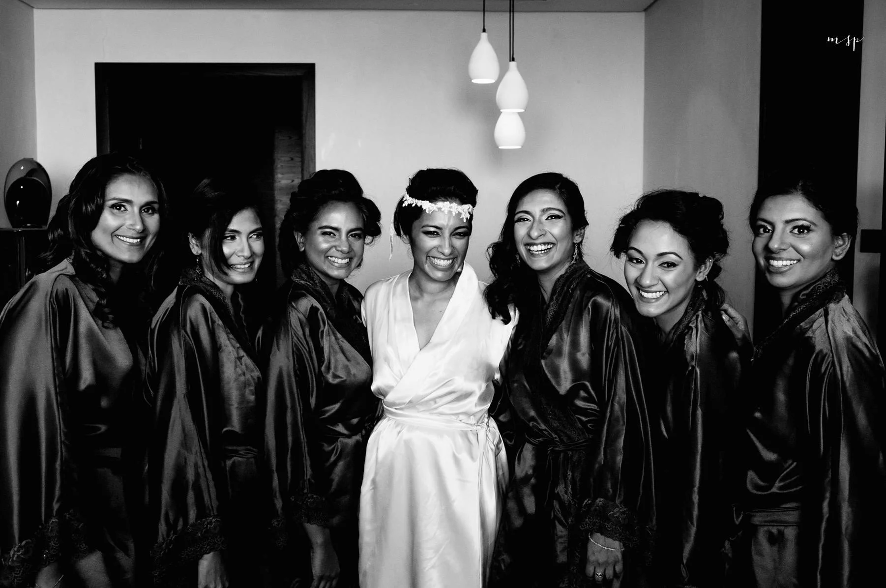 Group of seven smiling women in satin robes, with one dressed in a white robe and floral headband, standing indoors under hanging pendant lights.