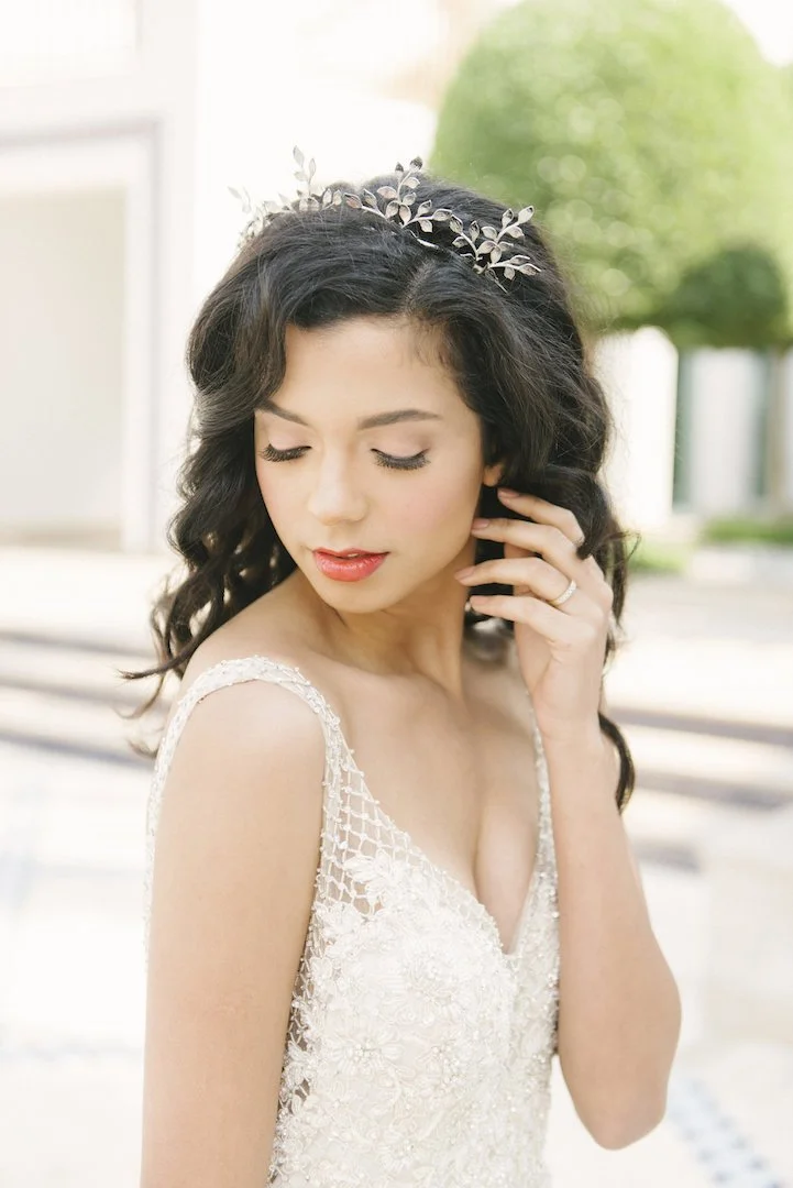 A woman with dark curly hair wearing a white lace dress with thin straps and a silver floral headband, posing outdoors with greenery in the background.