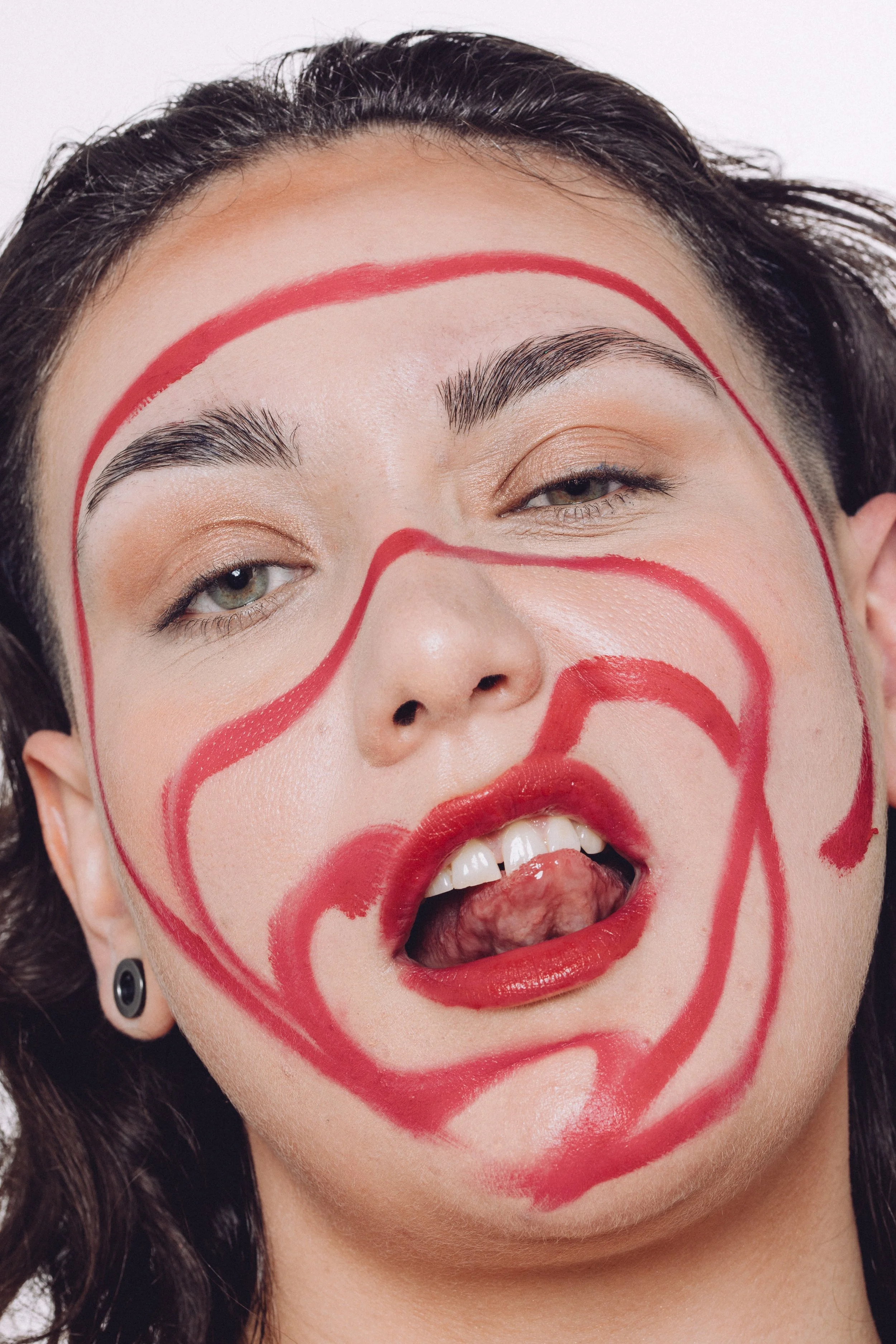 Close-up of a woman's face with red lines and shapes drawn on her face, one eye partially closed, mouth open with tongue sticking out.