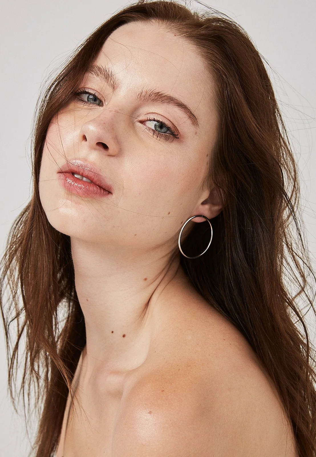 Close-up portrait of a woman with fair skin, wavy brown hair, blue eyes, wearing a silver hoop earring, with a neutral expression, and a plain light background.