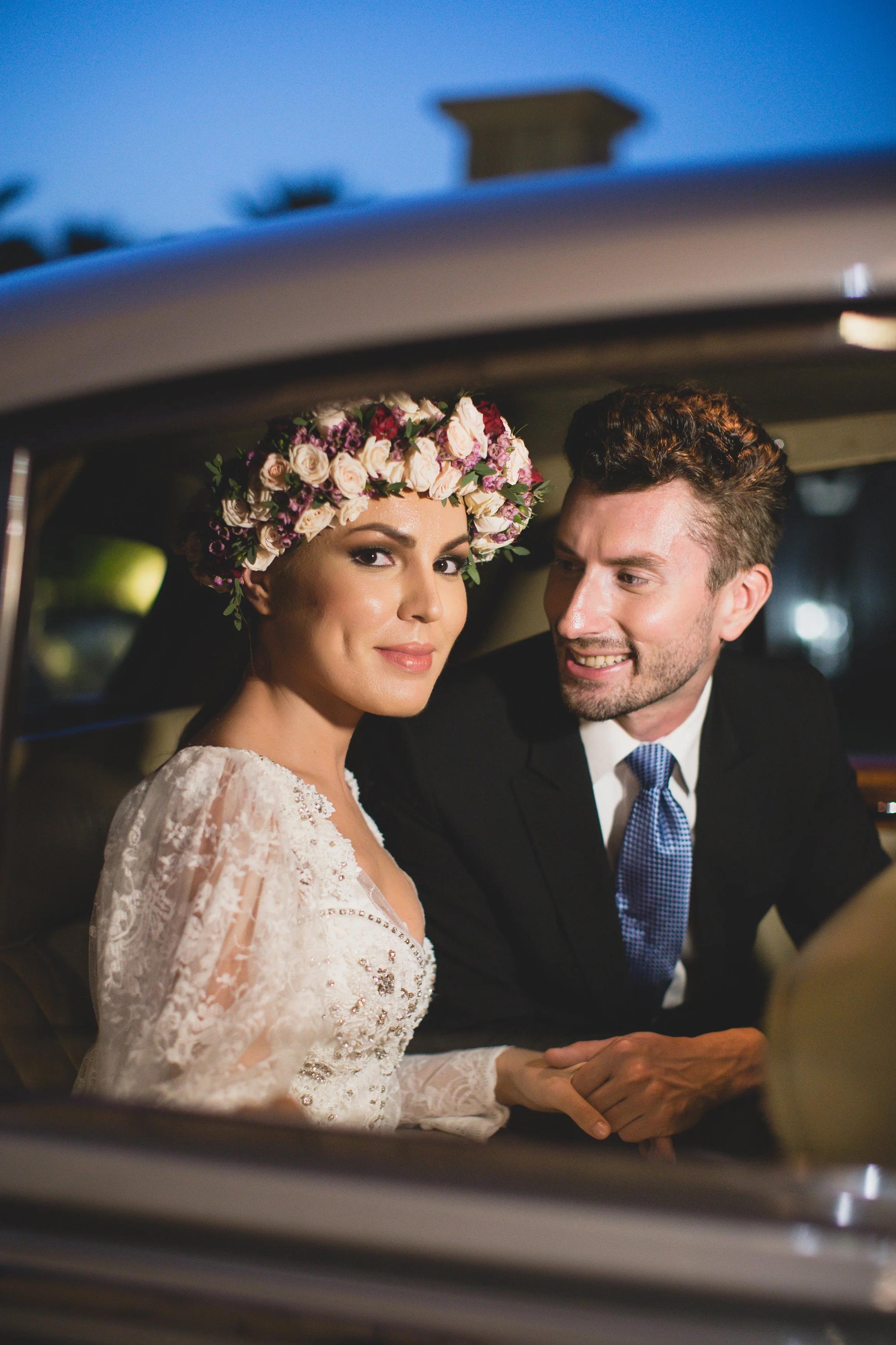 A bride and groom sitting inside a classic vintage car, with the bride wearing a floral crown and a lace wedding dress, and the groom in a dark suit with a blue tie, smiling at each other as they prepare for their wedding.
