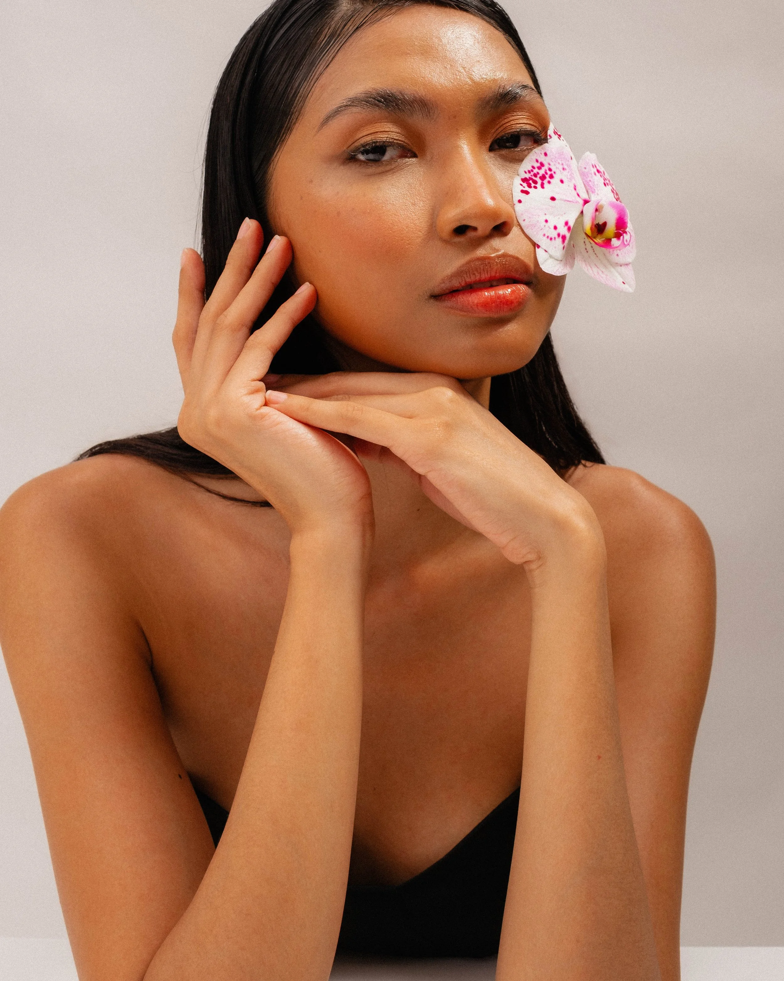 A woman with long dark hair and smooth skin, with a pink and white orchid flower near her right eye, posing with her hands gently touching her face against a plain light background.