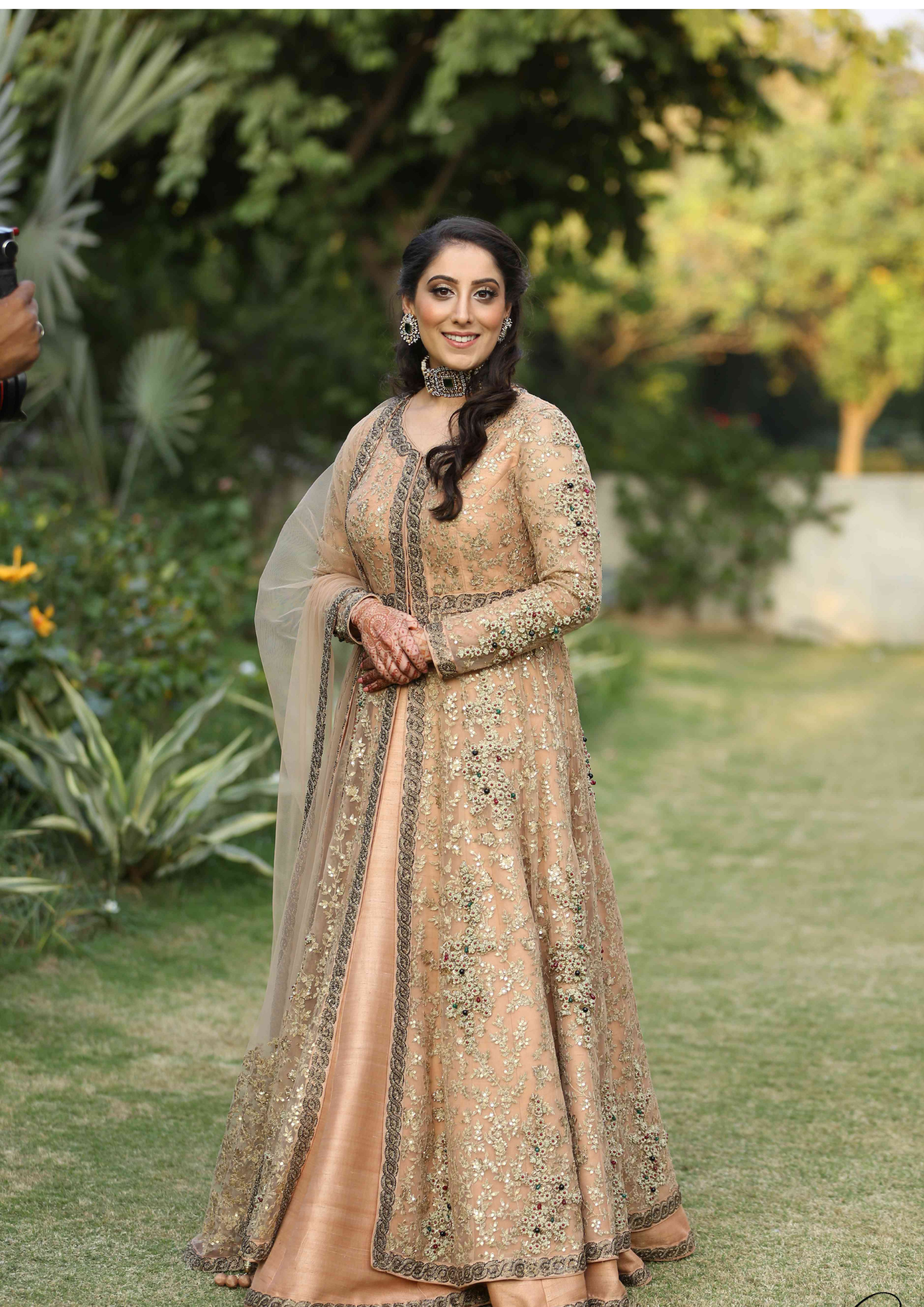 A woman in an elegant gold embroidered traditional dress, standing outdoors in a garden with greenery, smiling at the camera.