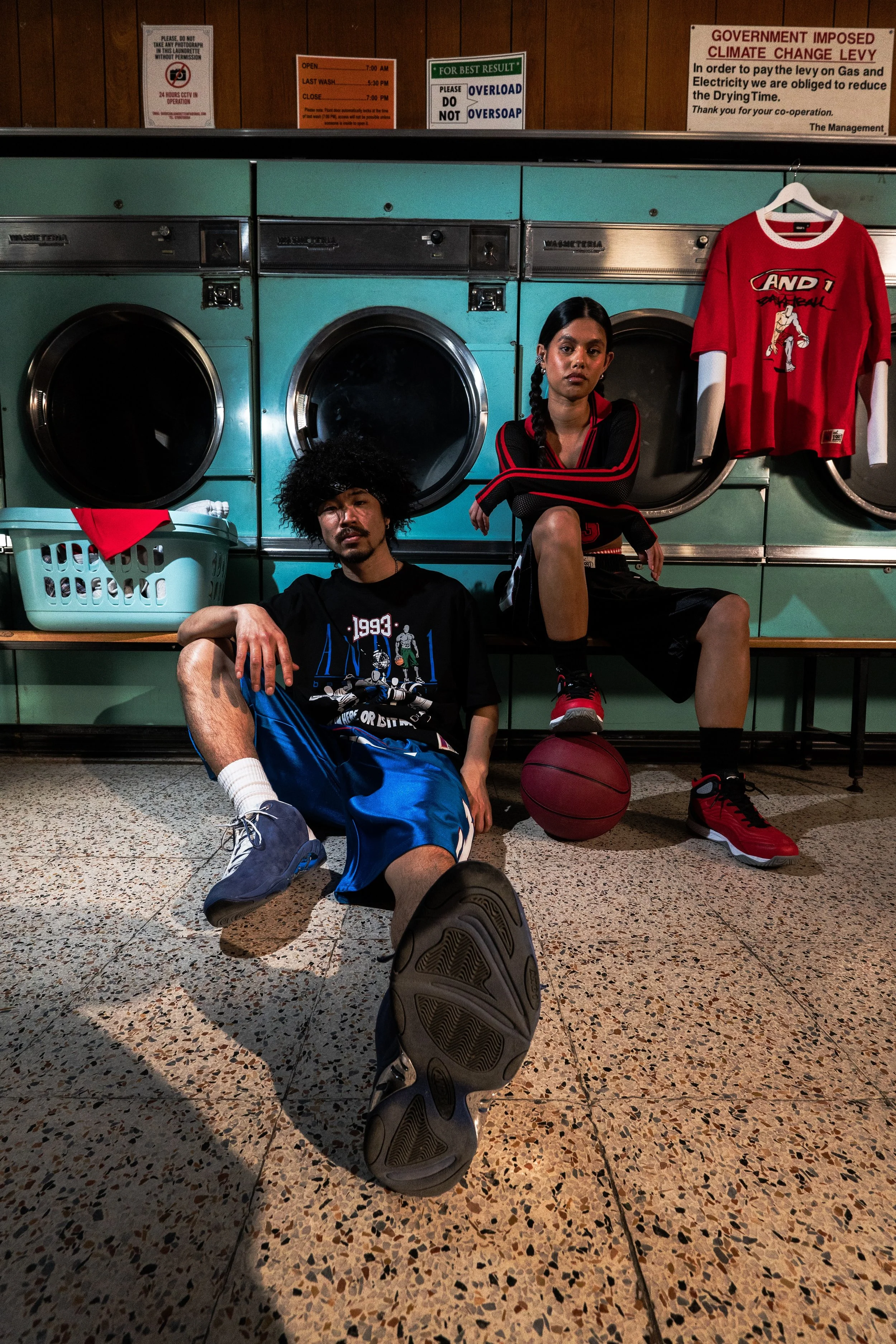 Two young adults in a laundromat with washing machines behind them. One is sitting on the floor with a sneaker closer to the camera, the other is sitting on a bench with a basketball. A red sports shirt is hanging on the wall.