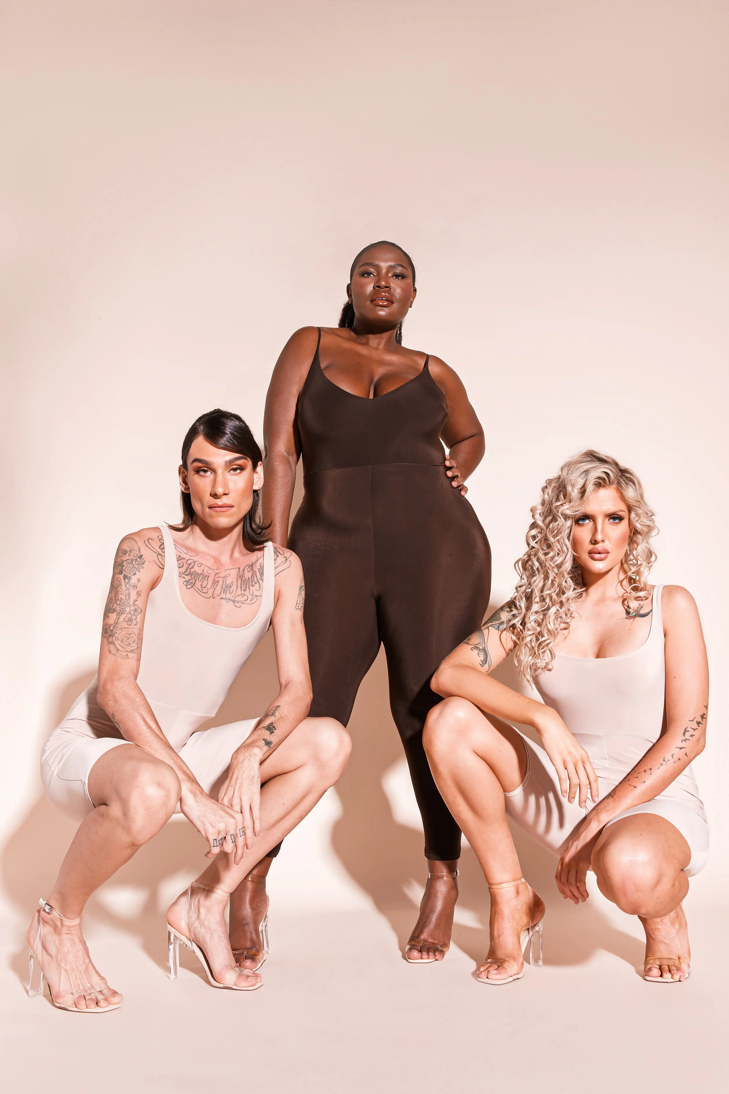 Three women with tattoos, posing in neutral-toned clothing and clear high-heeled shoes against a beige background.