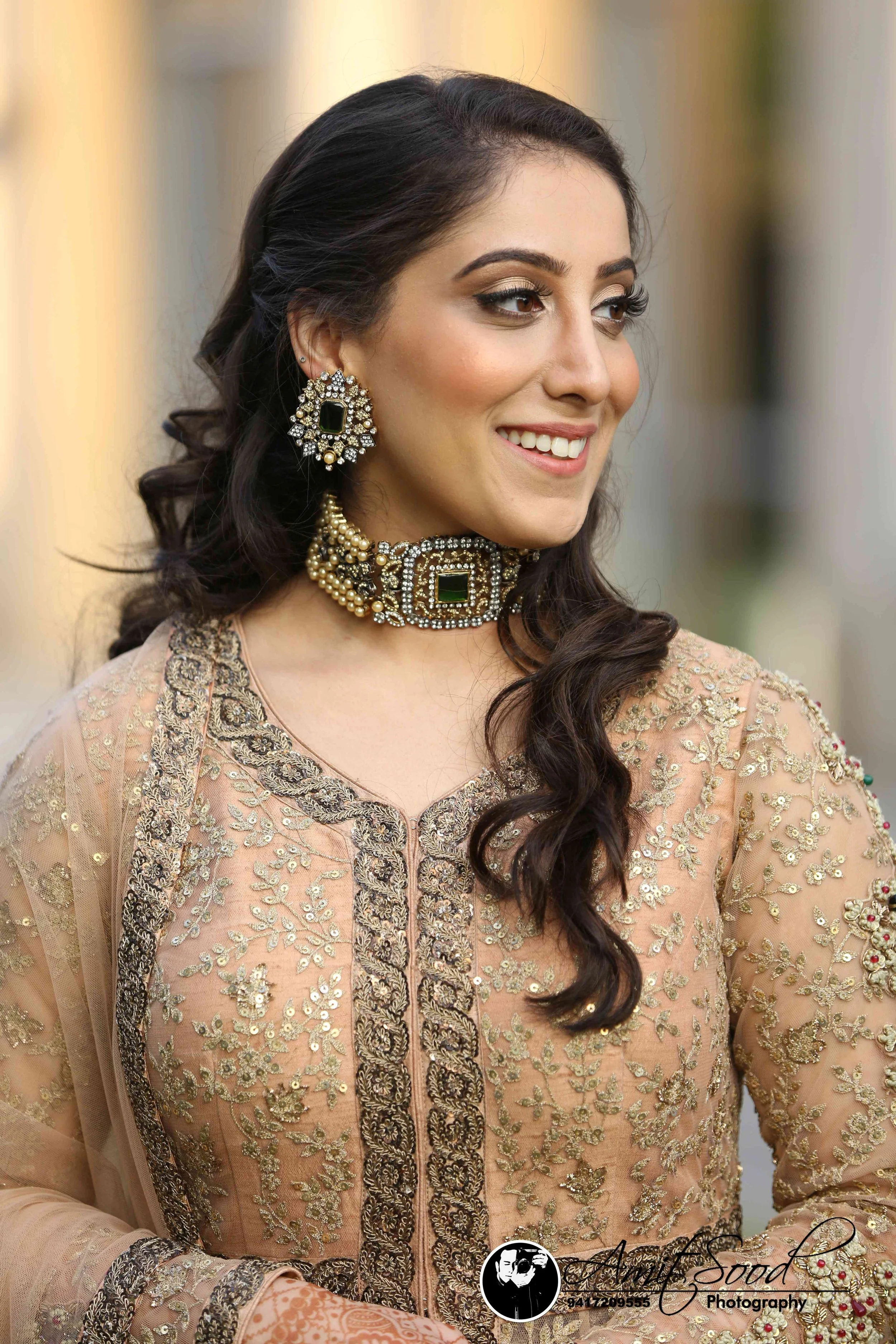 A woman in traditional Indian attire with intricate gold embroidery, wearing large jewelry including earrings and a choker, smiling and looking to her left.