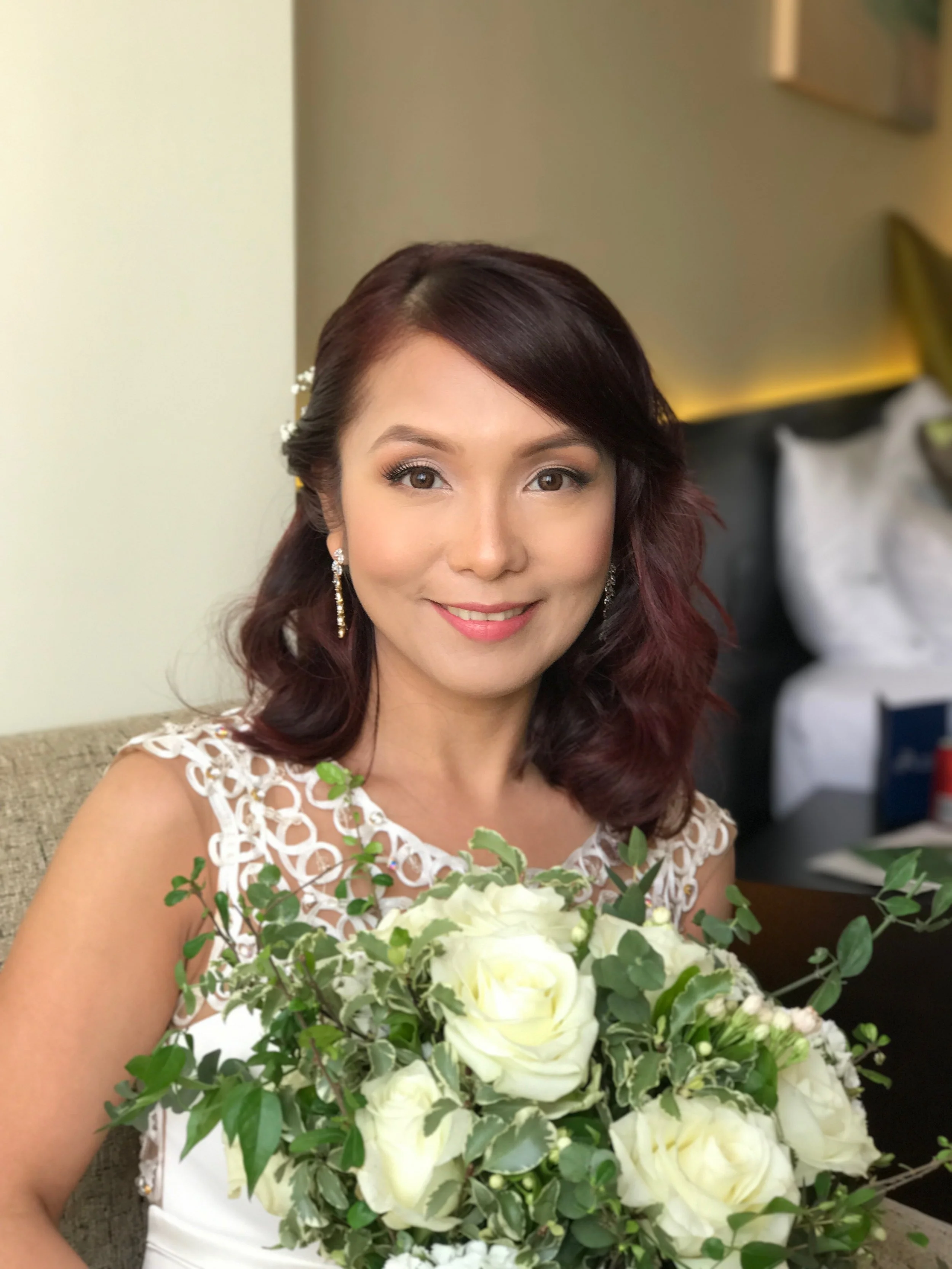 A woman with dark brown wavy hair holding a bouquet of white and green flowers, smiling, dressed in a white dress with lace details, sitting in a room with soft lighting.