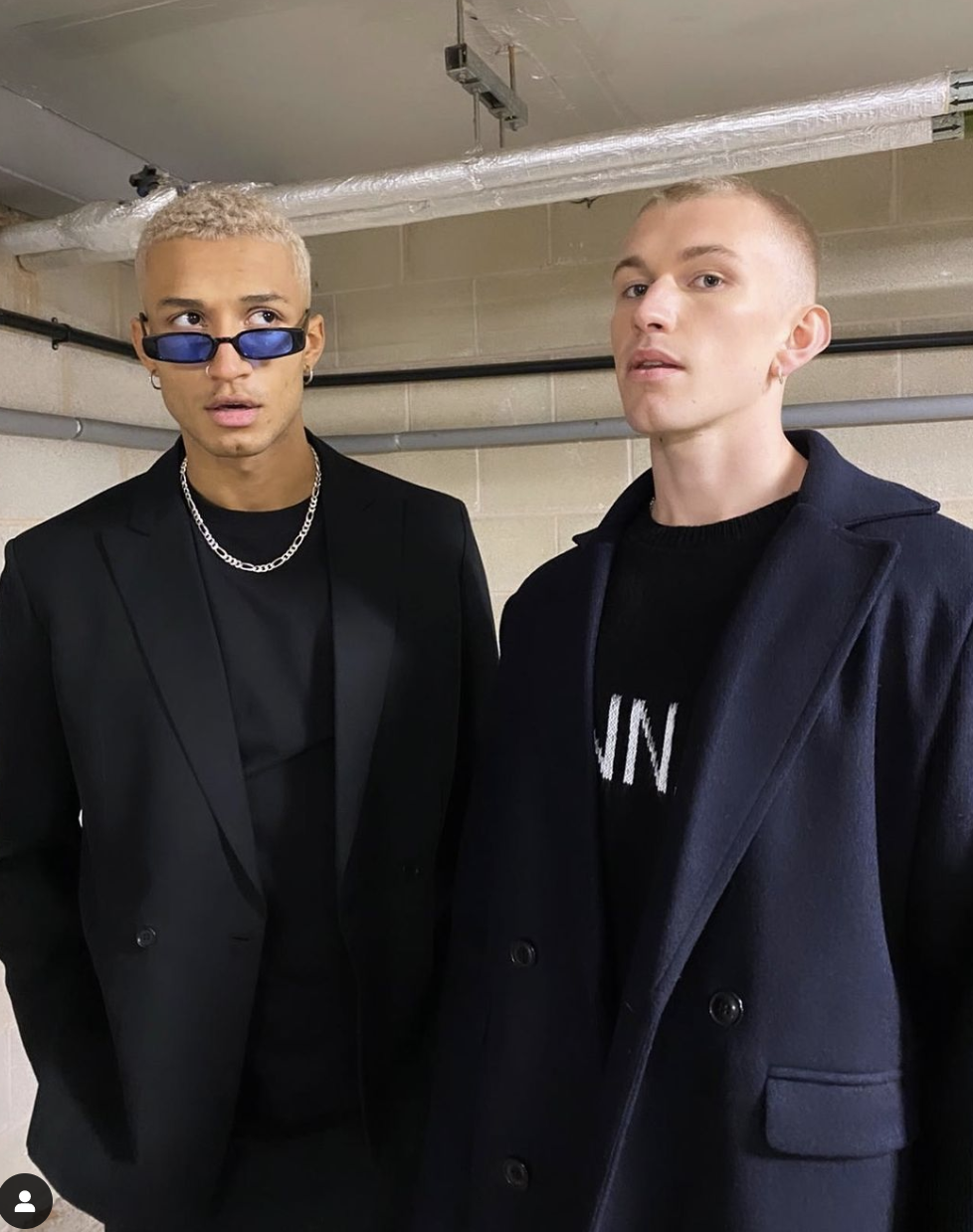 Two young men stand side by side in a doorway, one with platinum blonde hair, wearing dark sunglasses, a black blazer with satin lapels, and a silver chain necklace. The other has short, light hair, and is dressed in a dark coat over a black shirt. B