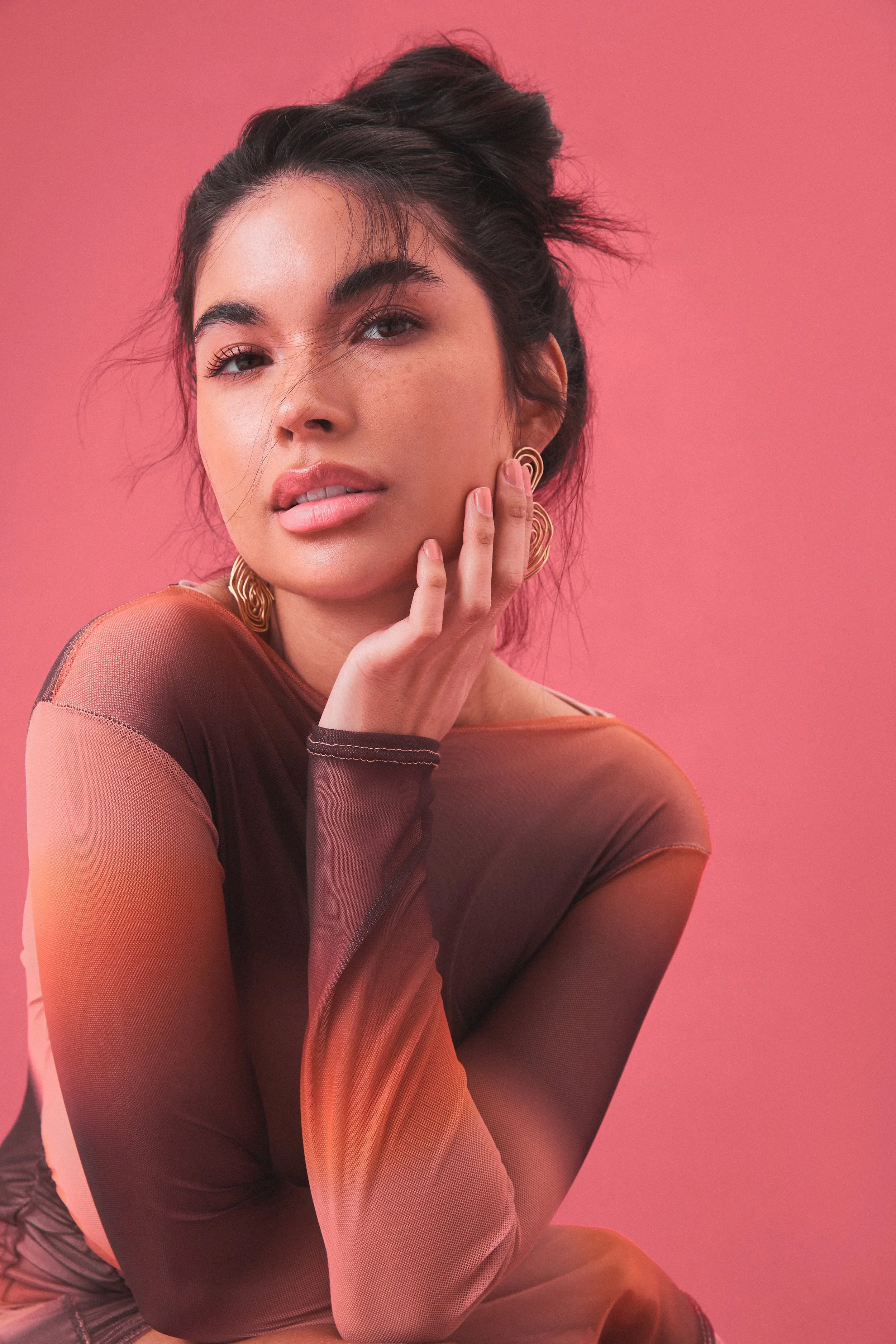 A young woman with dark hair styled in a messy bun, wearing gold earrings and a sheer, gradient long-sleeve top, sitting against a pink background, touching her face with her left hand and looking at the camera.