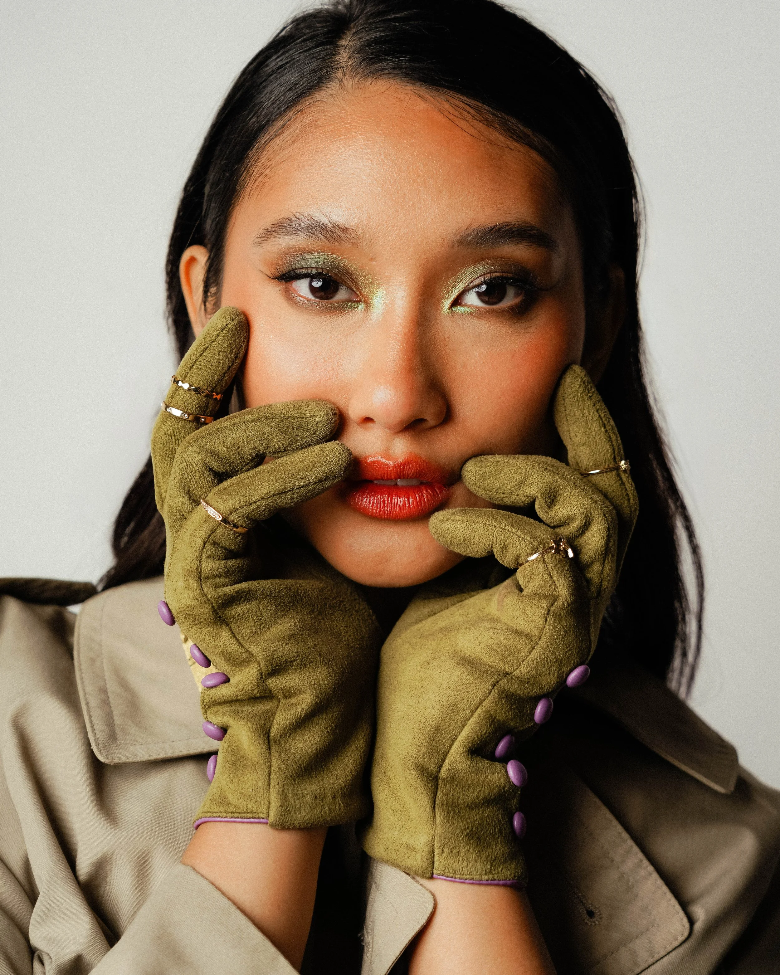 A woman with black hair and brown eyes wearing green gloves with purple accents, orange lipstick, and green eye makeup. She is posing with her hands framing her face, wearing multiple rings, and dressed in a beige trench coat against a plain backgrou