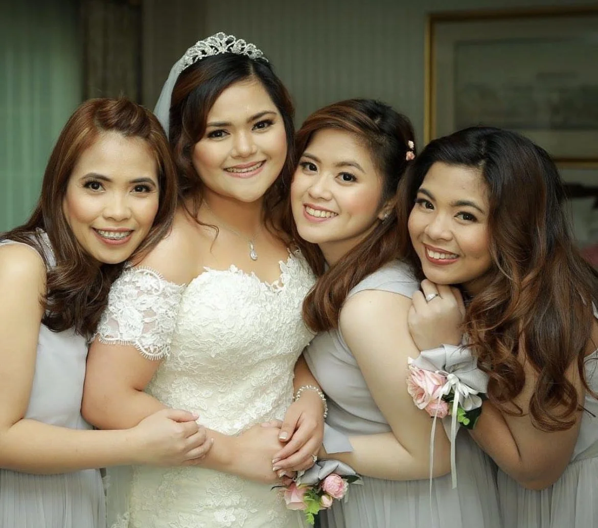 Group of four women at a wedding, with the bride in a white lace wedding dress and tiara, all smiling and holding hands.