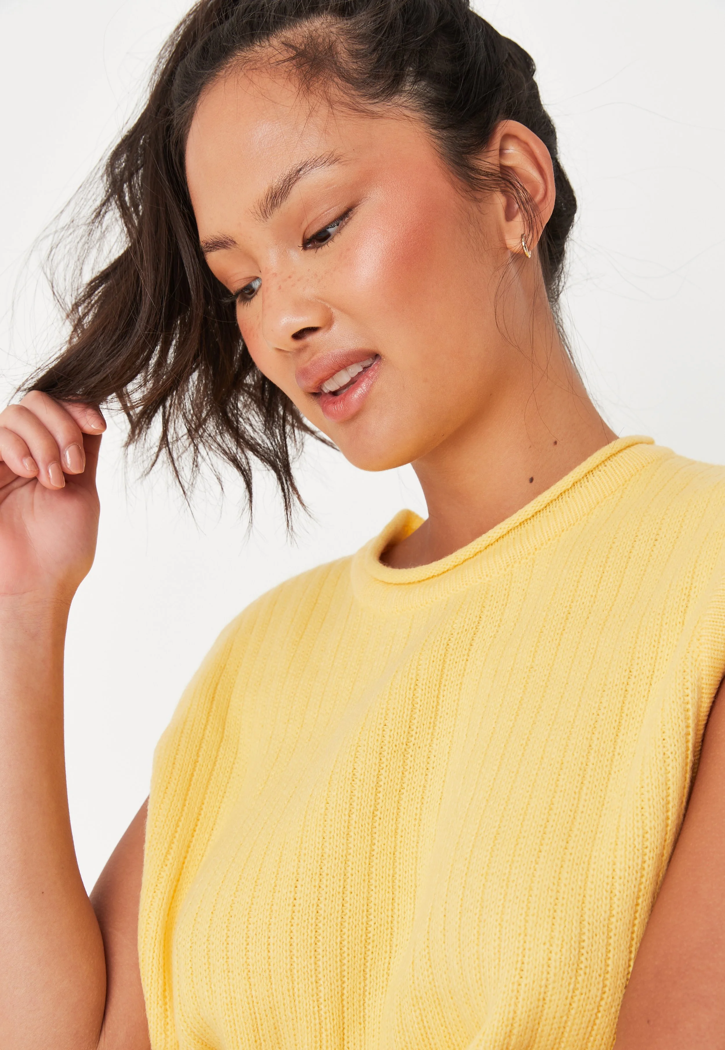 A young woman with short wavy hair wearing a yellow sleeveless top, looking down and touching her hair.