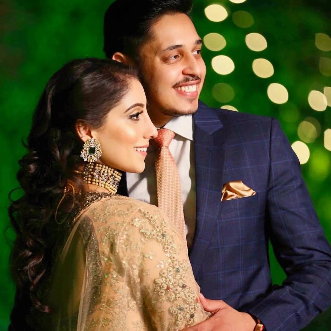 A smiling couple dressed in formal attire, standing closely against a green blurred background with lights, celebrating a special occasion or event.