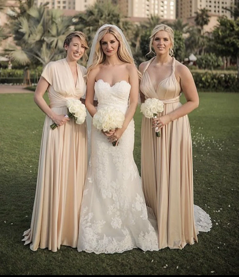 A bride in a white wedding dress with floral lace details, flanked by two women in matching blush-colored bridesmaid dresses, all holding white bouquets, standing outdoors on a grassy area with trees and buildings in the background.