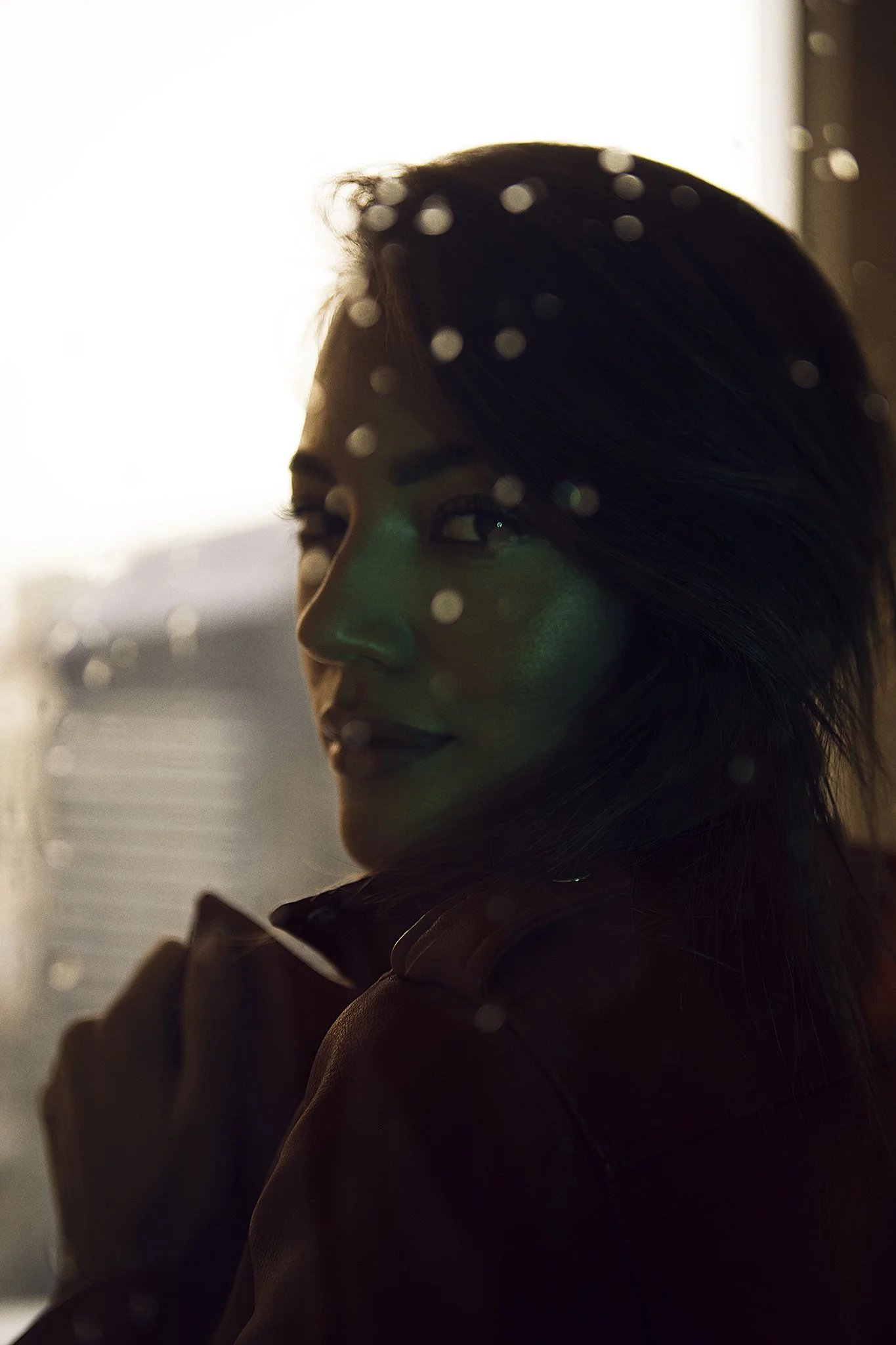 A woman with dark hair looking over her shoulder, illuminated by warm backlighting, with water droplets on a glass surface in front of her.