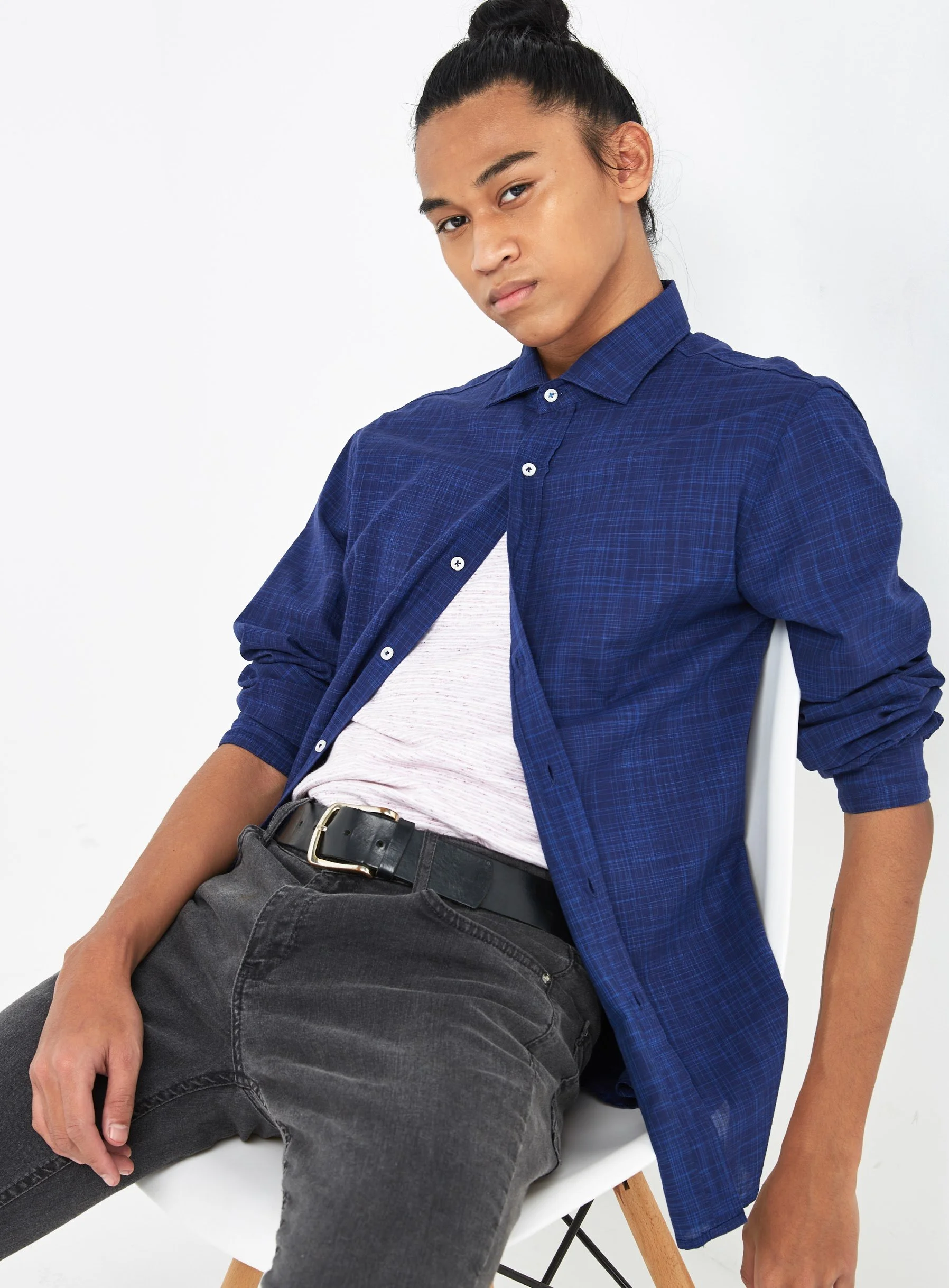 Young man sitting on a white chair, wearing a blue button-up shirt over a white t-shirt, black jeans, and a black belt, with his hair tied up in a bun, against a plain white background.