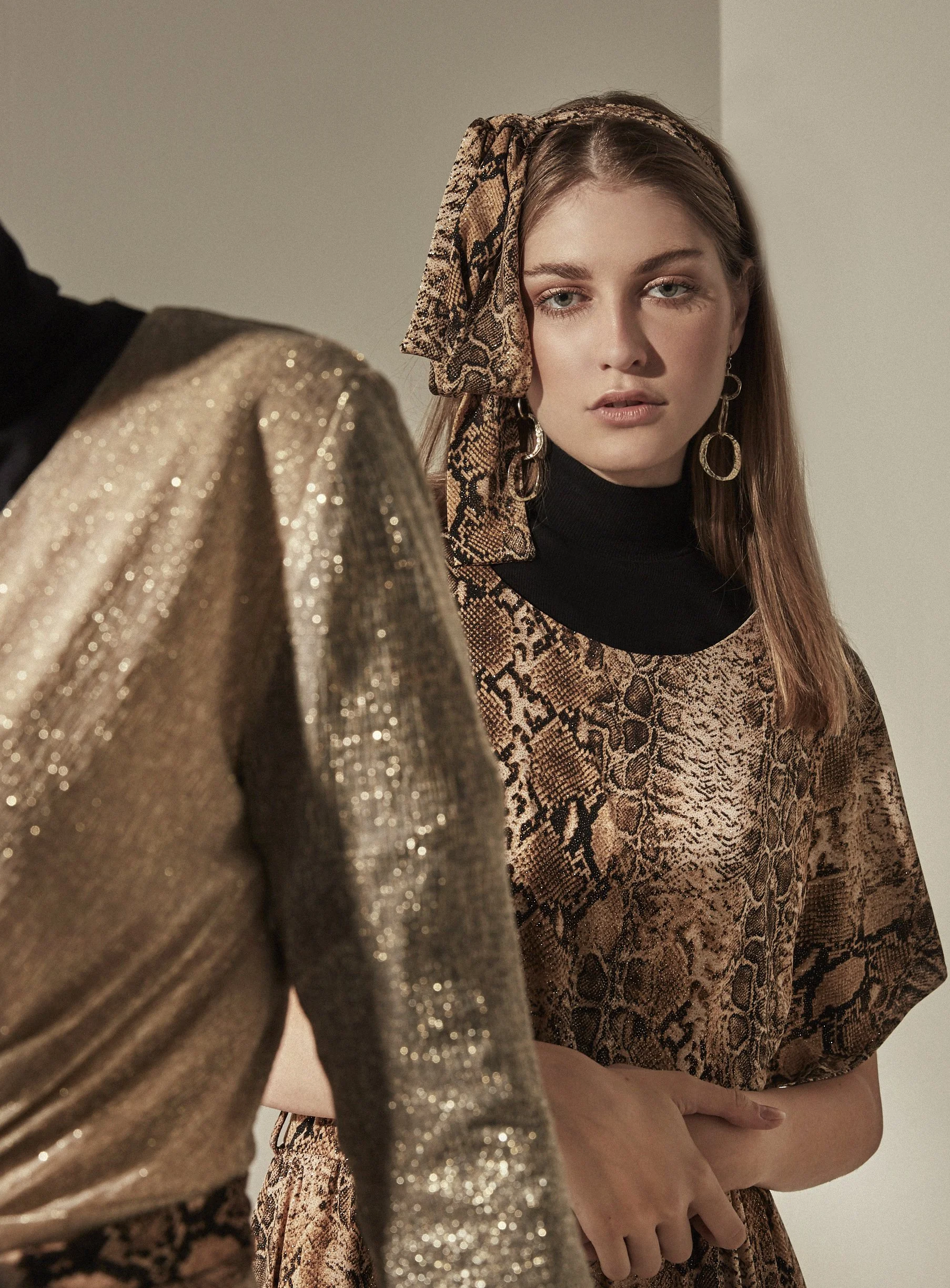 A woman wearing a snake print dress and headscarf, with a black turtleneck underneath. She has long hair, hoop earrings, and a serious expression.