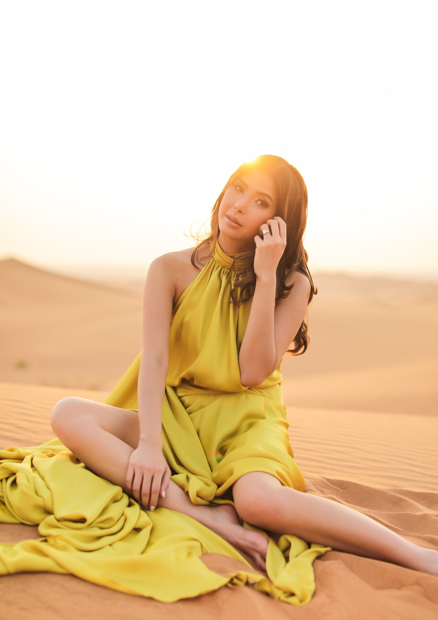 A woman sitting on desert sand dunes at sunset, wearing a yellow dress, with the sun shining behind her head.