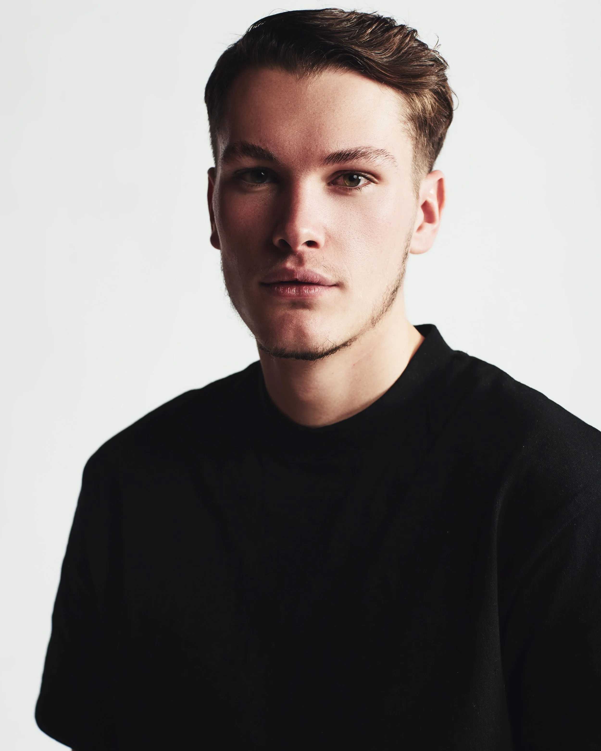 Portrait of a young man with short, styled brown hair, light skin, wearing a black shirt, against a plain white background.