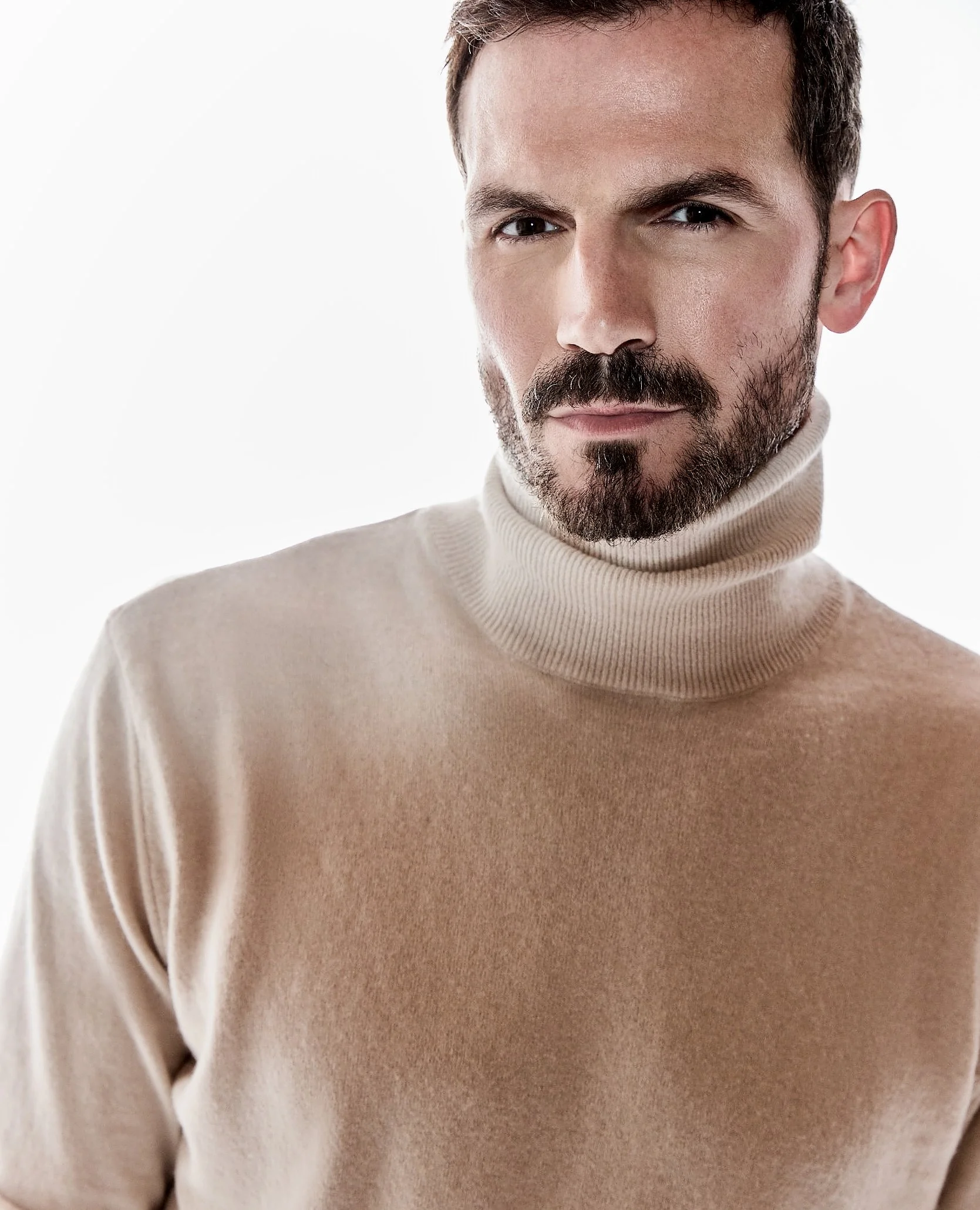 A man with short brown hair, a beard, and mustache wearing a beige turtleneck sweater, looking at the camera with a neutral expression.