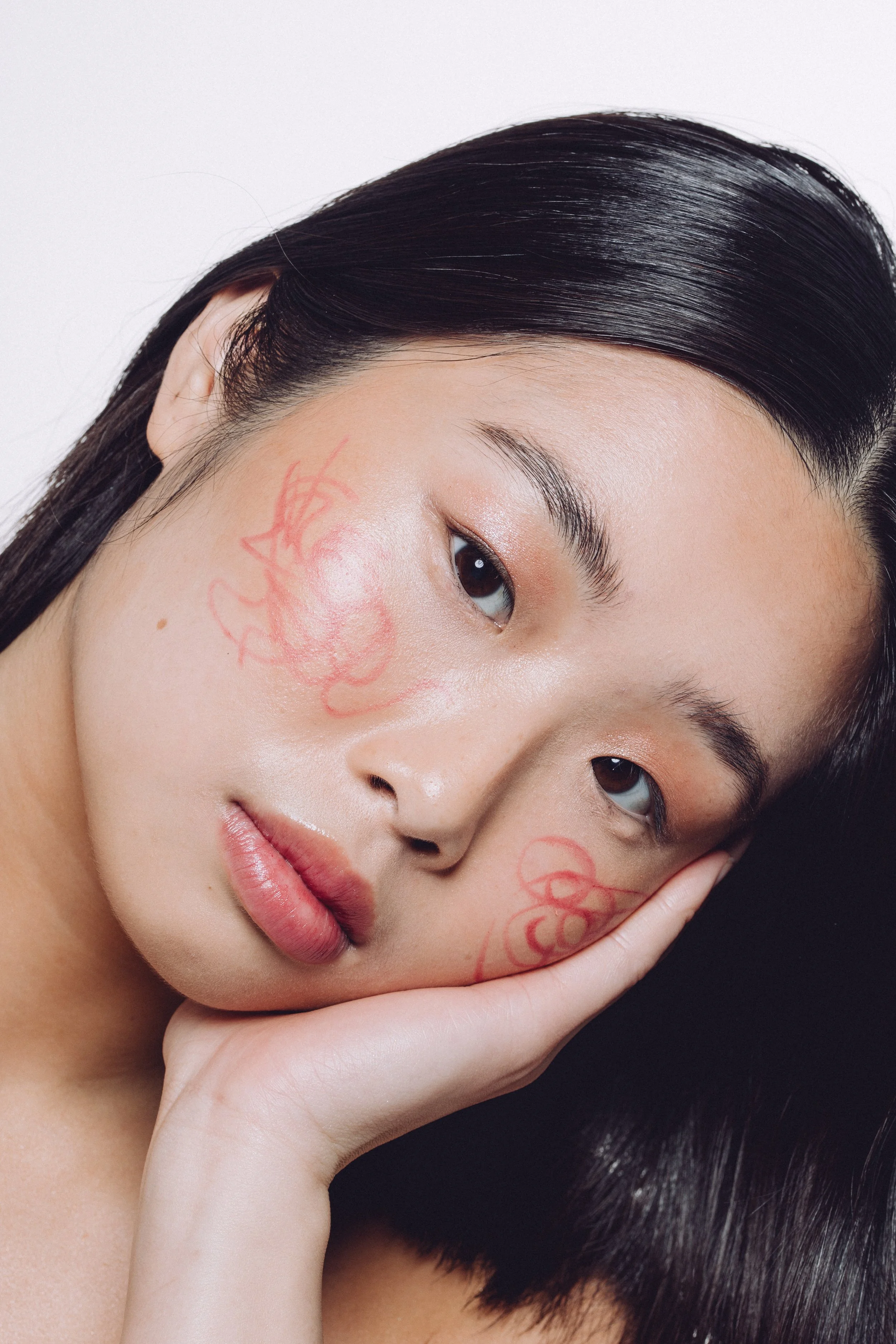 Close-up portrait of a young woman with dark hair resting her face on her hand, with red temporary tattoos on her cheek.