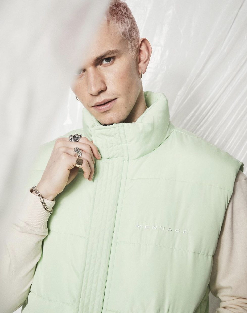 A young man with pink hair and earrings wearing a light green padded vest over a beige long sleeve shirt, holding his chin with his right hand, against a white background.