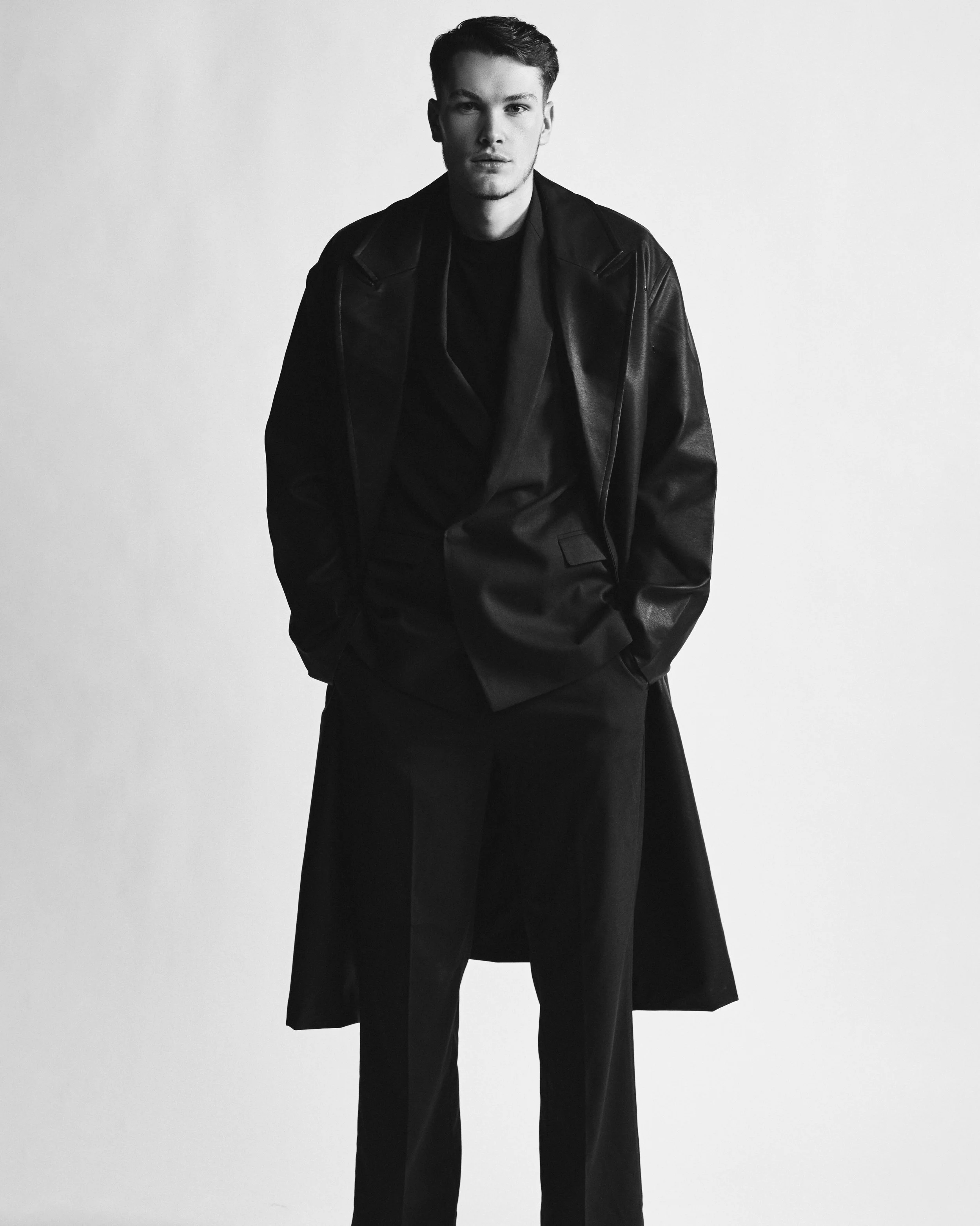 A young man in a black leather jacket and dark pants poses with his hands in his pockets against a plain light background.