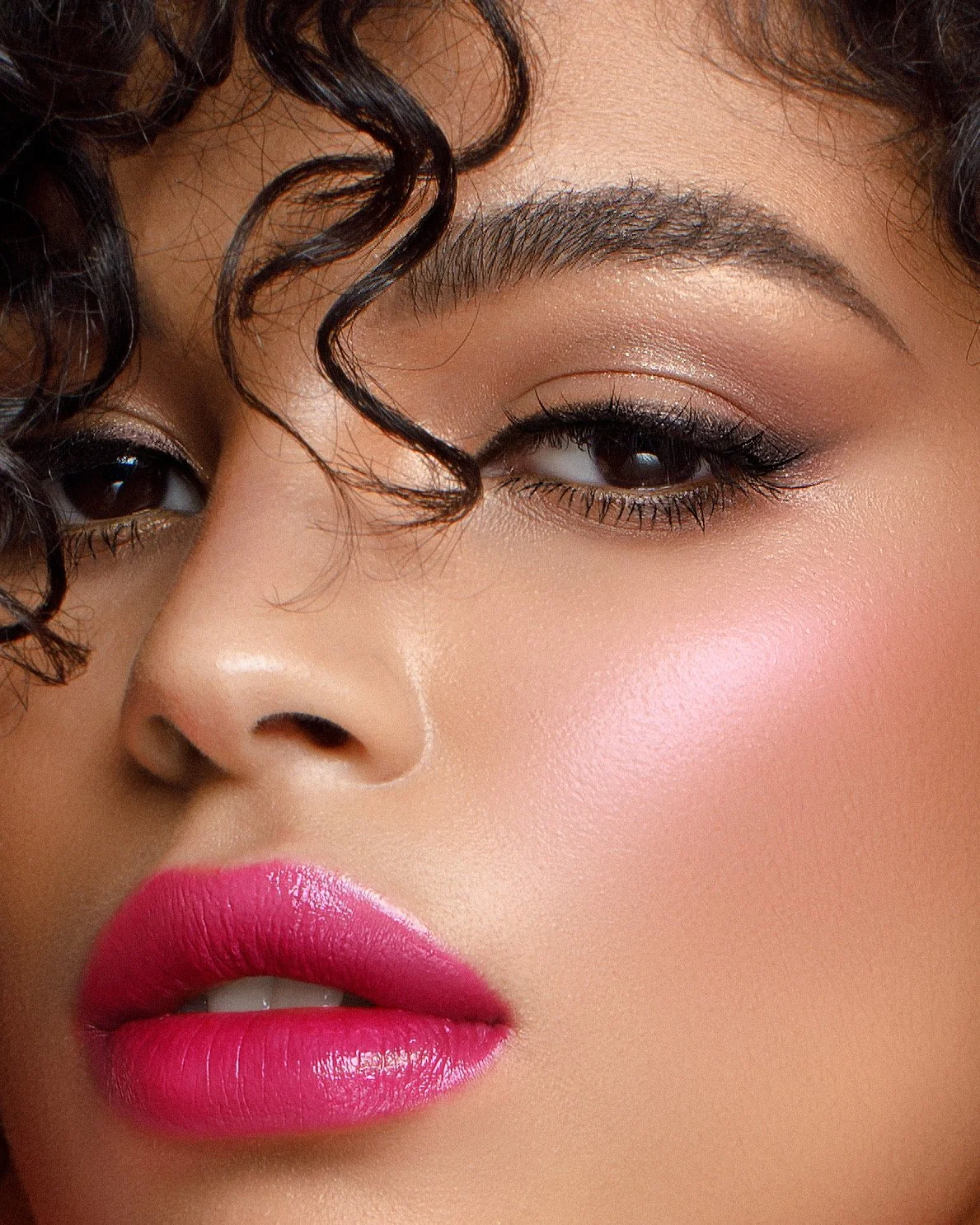 Close-up of a woman's face showing her eye, nose, and lips. She has dark, curly hair and makeup including eyeshadow, eyeliner, mascara, and pink lipstick.