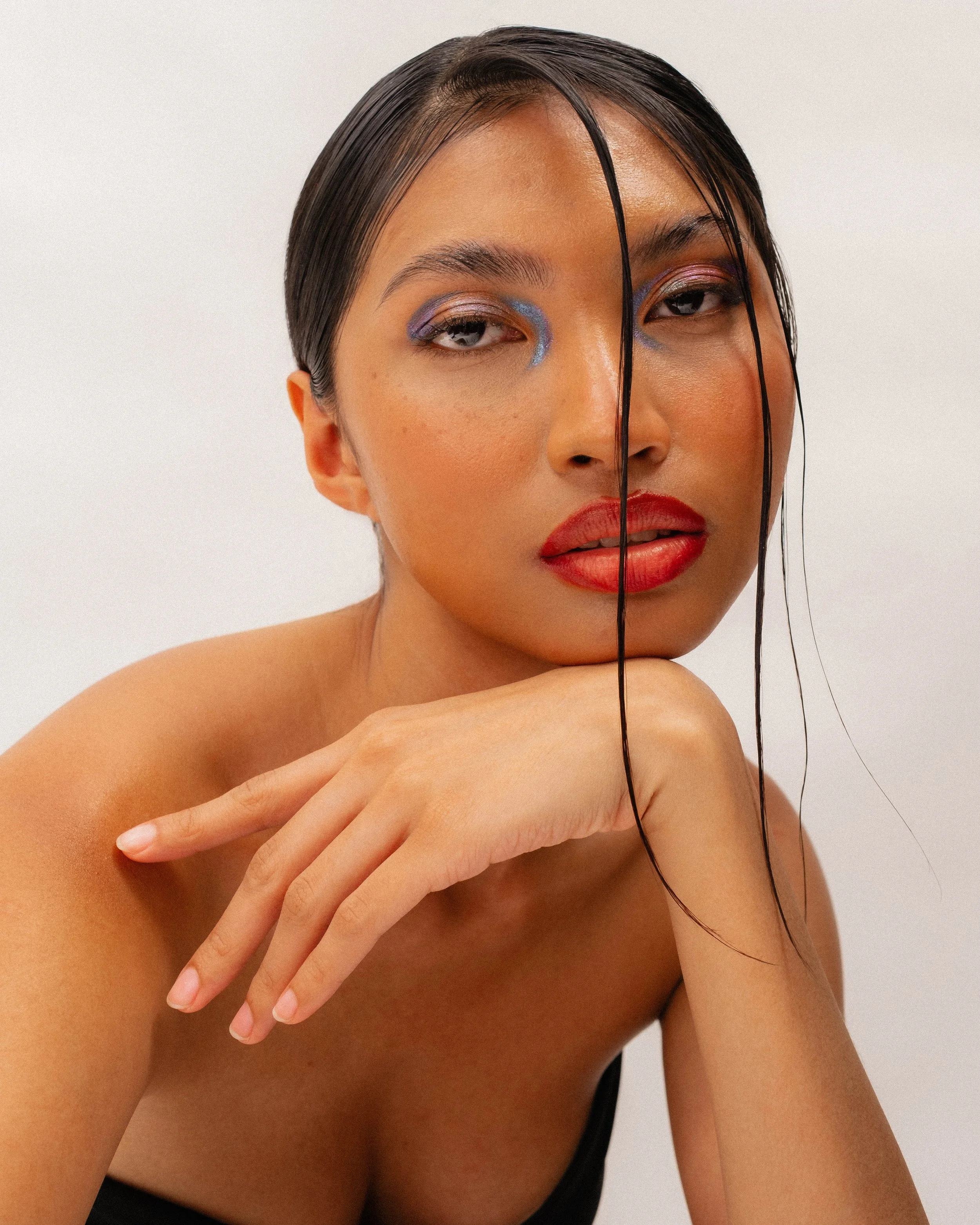 Close-up portrait of a woman with wet hair, wearing bold makeup with colorful eyeshadow and red lipstick, resting her chin on her hand.