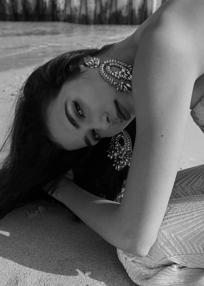 A woman lying on the beach, resting her head on her arm, wearing large, ornate earrings and a strapless top, with long dark hair spread out on the sand.