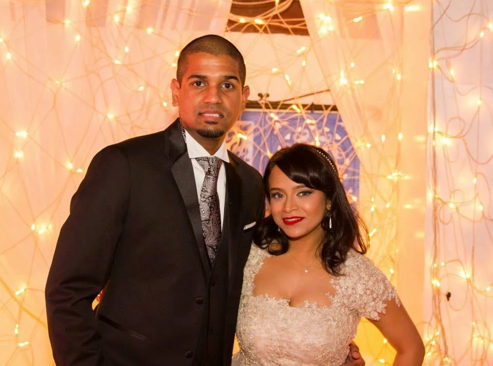 A man in a black suit and a woman in a light-colored dress posing together at a decorated event with warm string lights in the background.