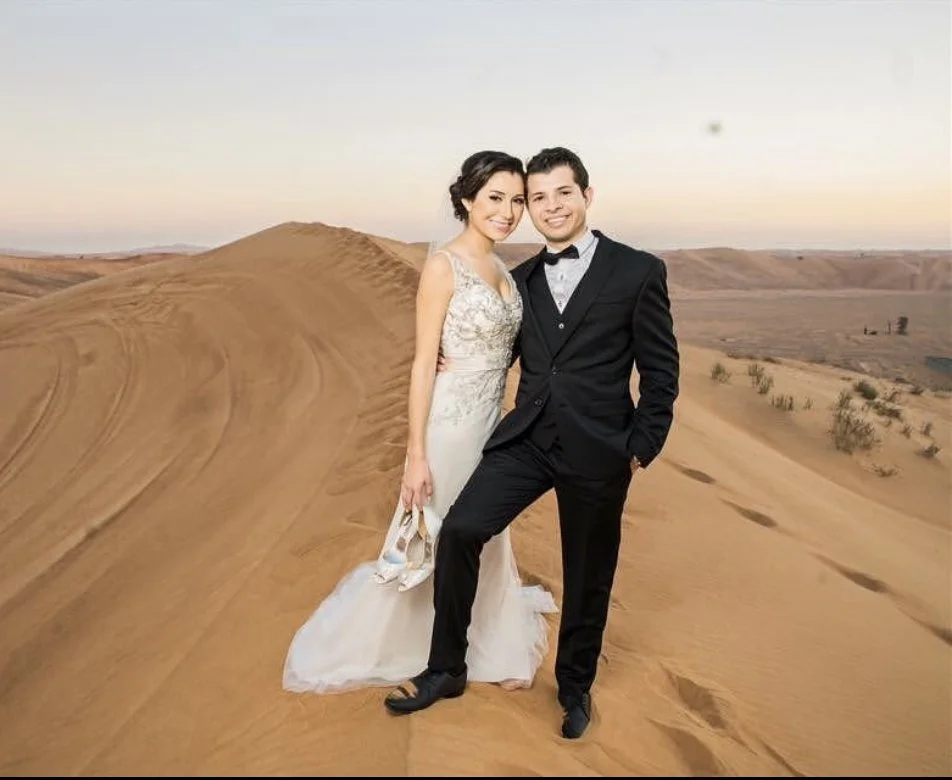 A couple in wedding attire standing on sand dunes in a desert at sunset, with sparse vegetation in the background.