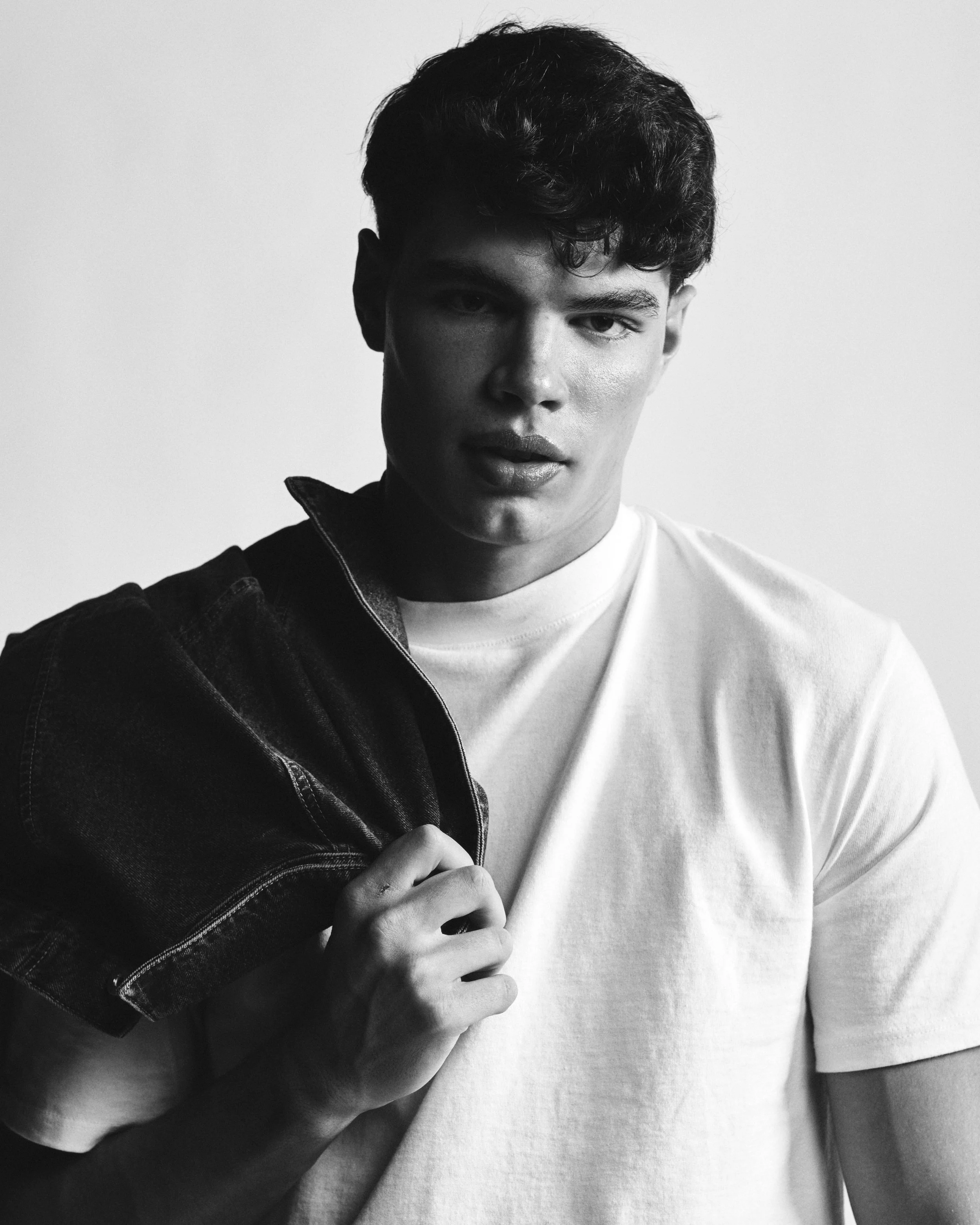 Black and white portrait of a young man holding a denim jacket over his shoulder, wearing a light t-shirt, with a serious expression.