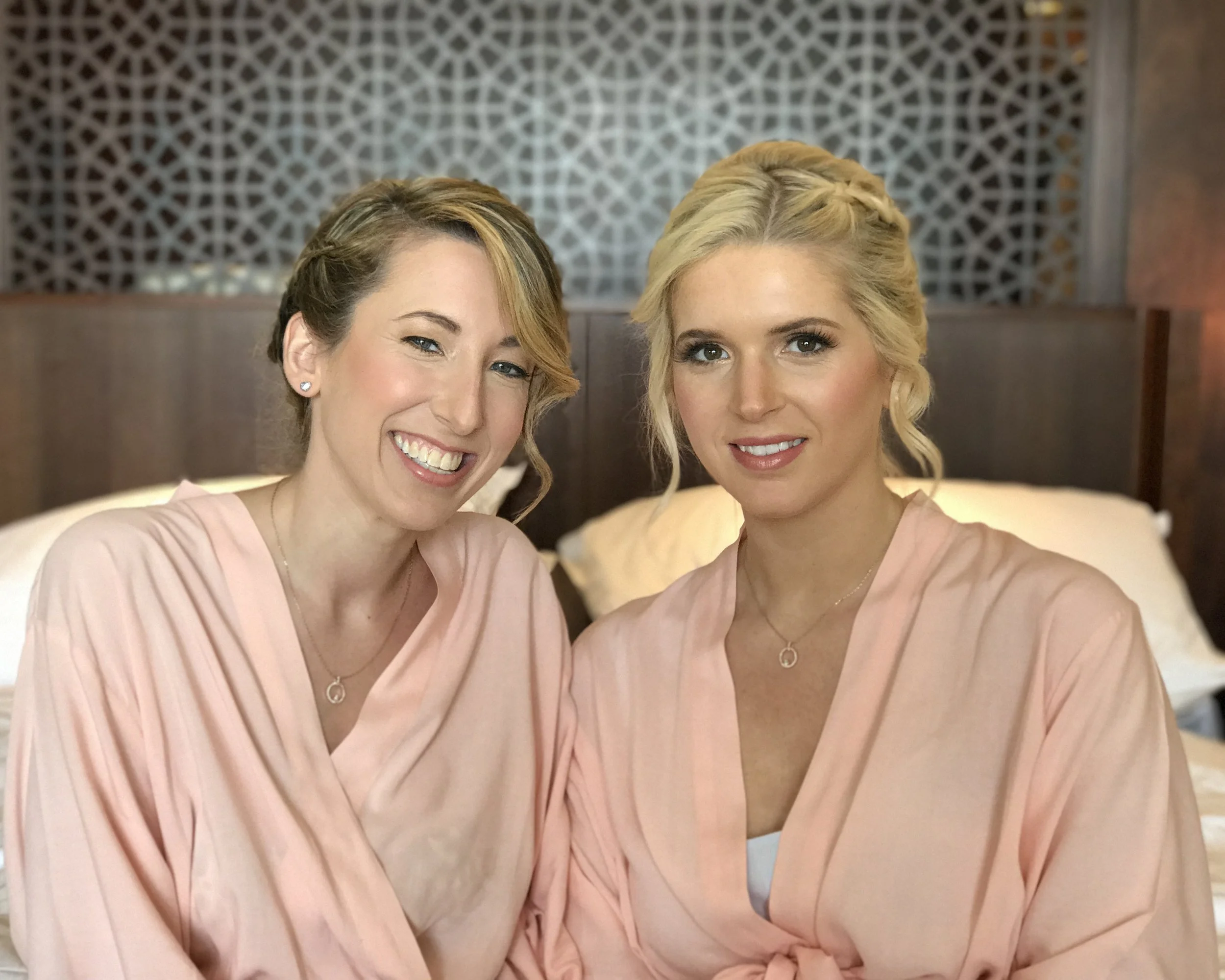 Two women in peach robes sitting on a bed, smiling at the camera, with a wooden headboard and decorative wall behind them.